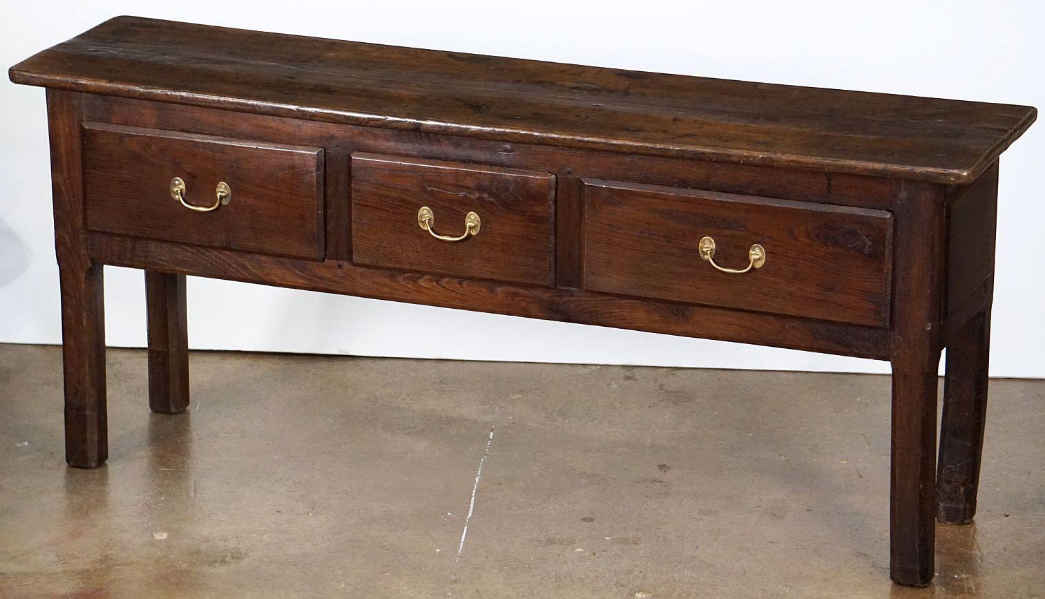 A handsome French console sideboard or serving table of patinated chestnut wood, featuring a plank top over a paneled frieze with three drawers, each with brass hardware pulls, and resting on four chamfered legs.

