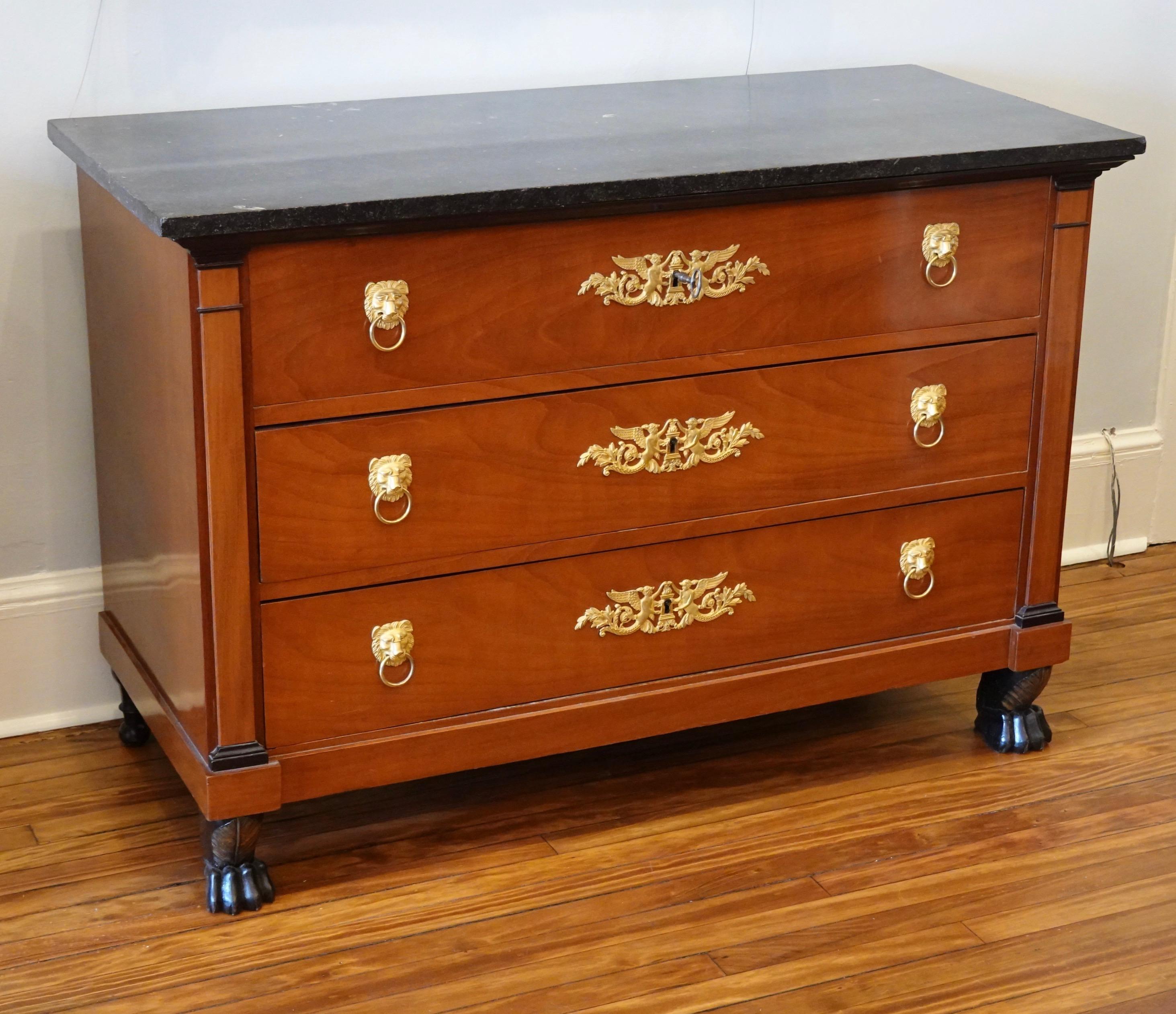 French Consulat period mahogany commode with three-drawers, ebonized griffin feet and pilasters, and black fossil marble top. Gilt bronze escutcheons in the form of winged angels and lions head pulls, working locks and key. Marble top appears