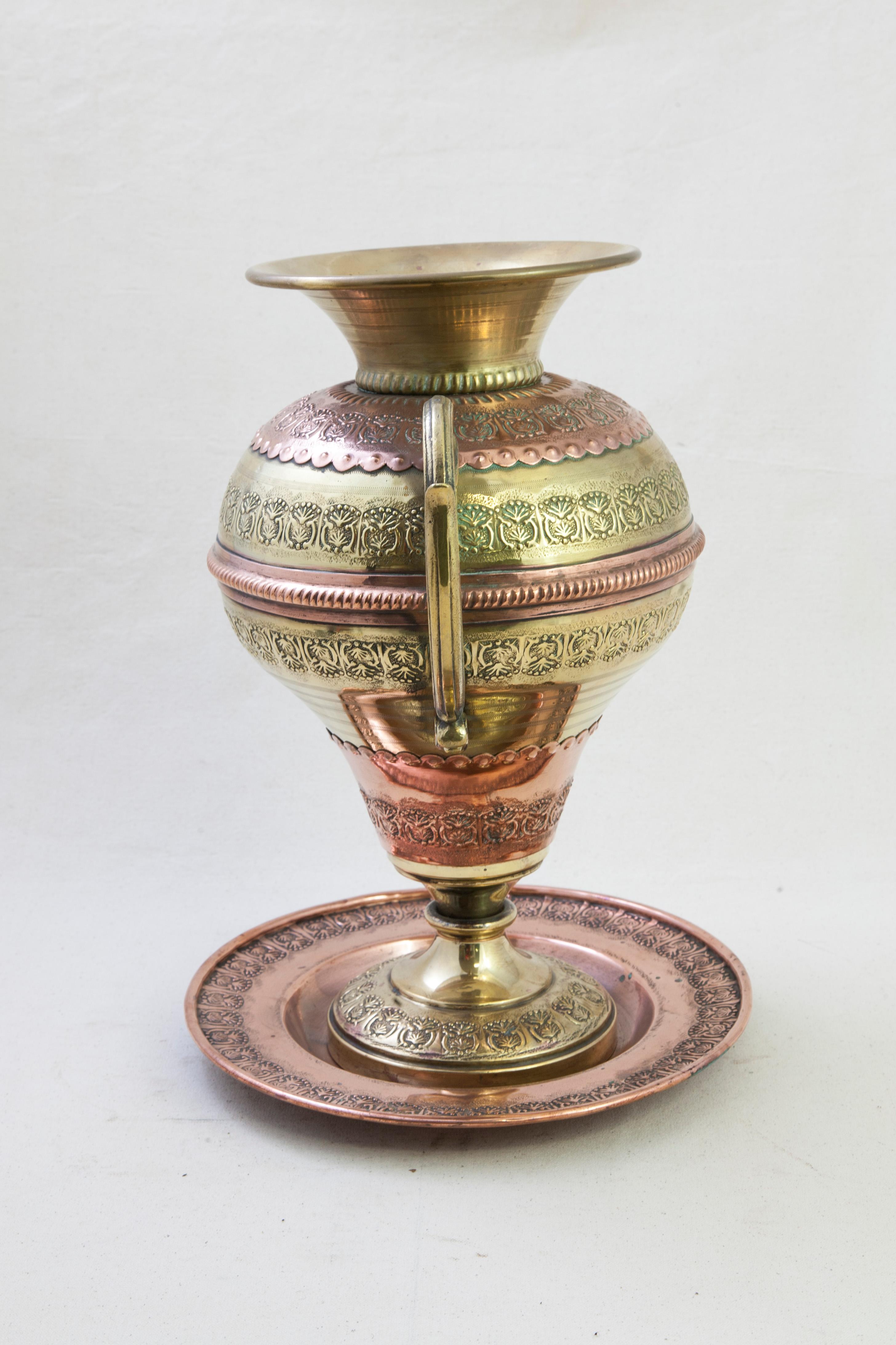 This turn of the twentieth century copper and brass urn or vase comes with its original platter. The vase is detailed with repousse leaf and scrolling patterns alternating with incising and scalloped edging. A scrolling leaf handle appoints each
