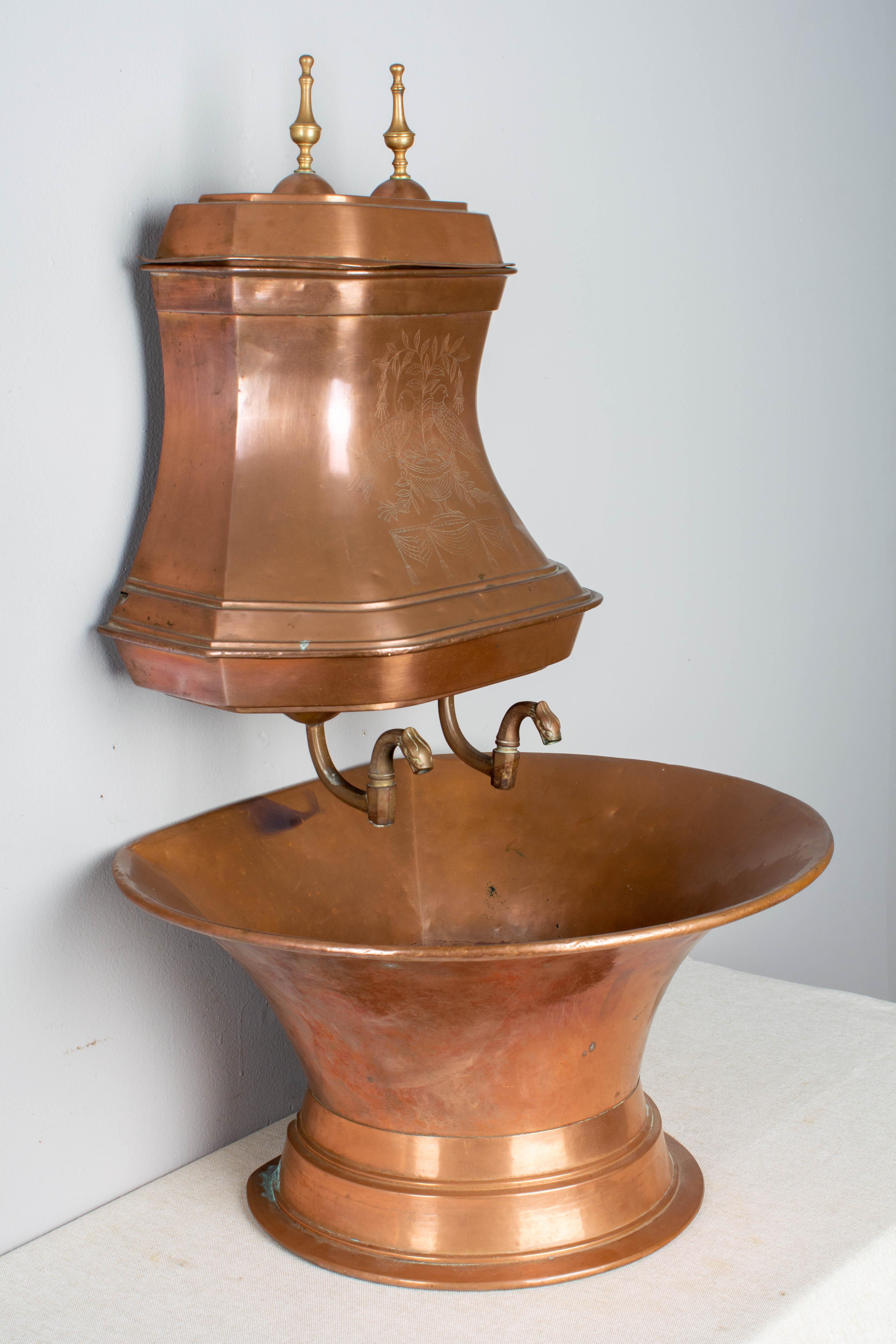 A French copper lavabo, or water reservoir with large basin. The tank has a repoussé decoration of birds and stylized flowers on the front and cast brass finials on the lid. The two cast brass water taps allow the water to empty into the large basin