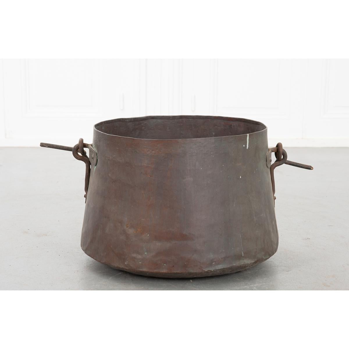 A sizable antique copper pot, equipped with a forged iron handle that can be used to suspend the vessel over a fire. The pot has a hammered exterior that gives the antique a styled finish. The copper has patinated wonderfully, with some verdigris