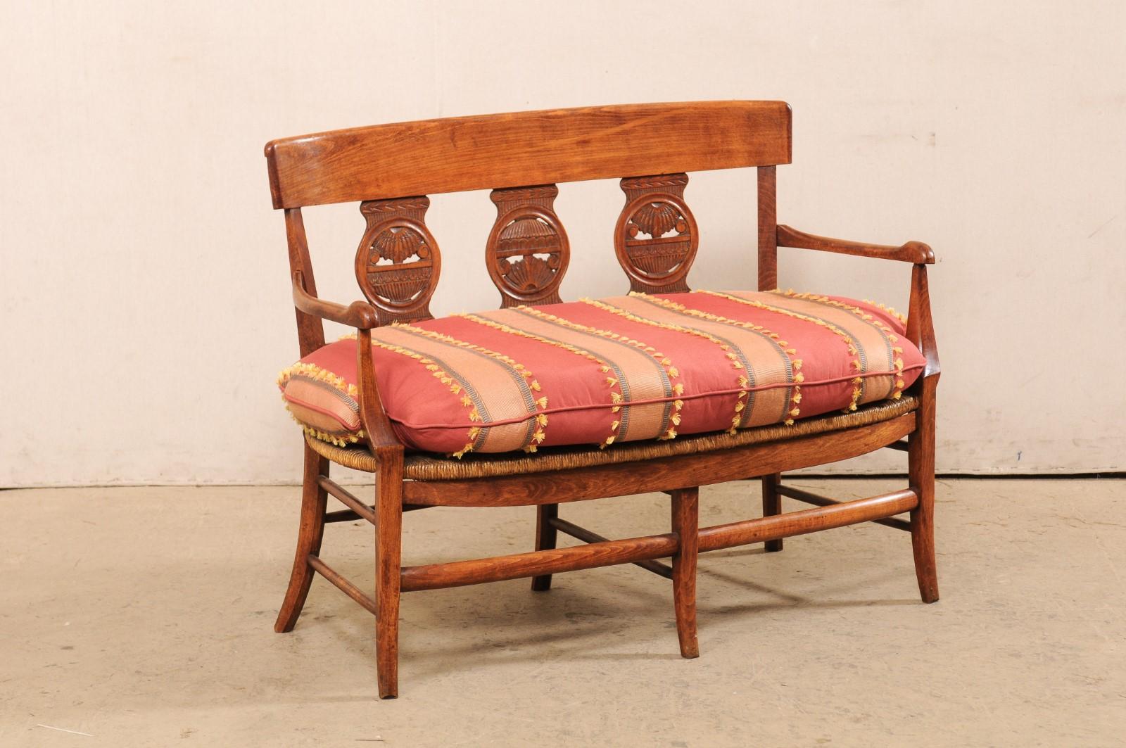 A French carved-wood loveseat with rushed seat from the early 19th century. This antique bench from France features three back-splats carved with circular centers and Neoclassic influences, set within a more French country frame consisting of clean,