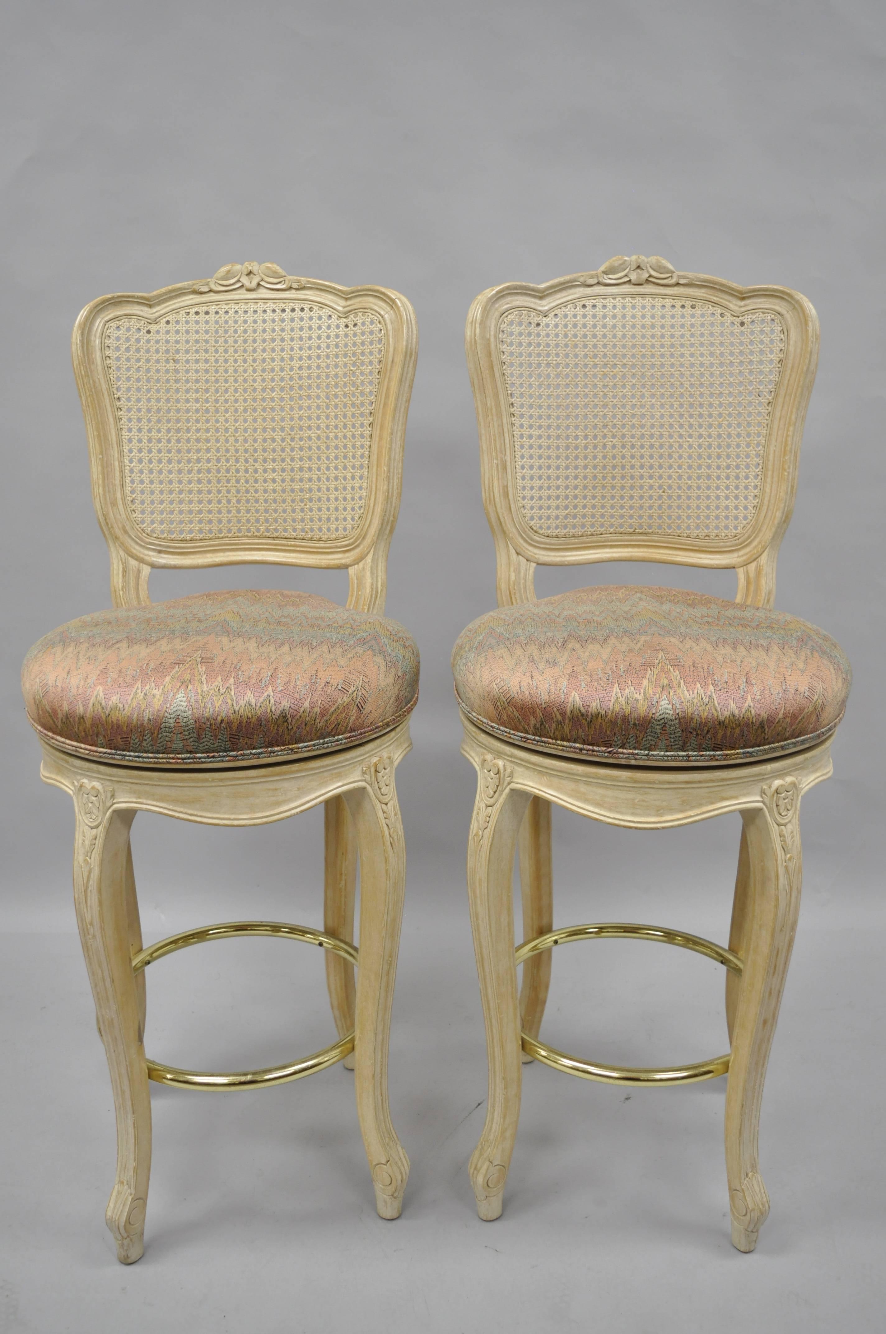 Pair of vintage French country Louis XV style cane back swivel bar stools. Stools feature solid carved wood frames, upholstered swivel seats, cane backs, cabriole legs, white washed distressed finish, brass footrest, classic French