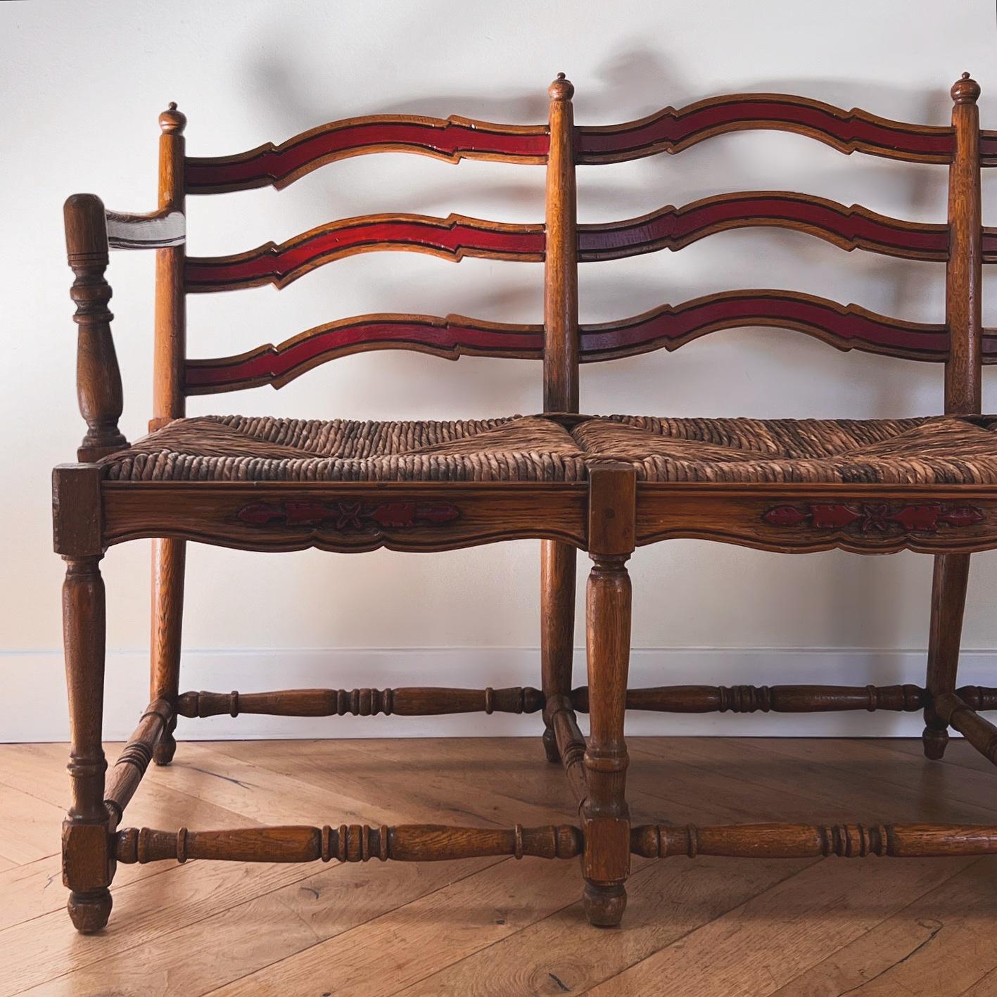 A wicker rush and oak wood ladder back Provençal painted bench, 20th century. Probably circa early 1950s. Featuring spindle and painted cerise detail. Fabulous indoors or as an outdoor patio bench - this piece would be lovely a bit weathered. For