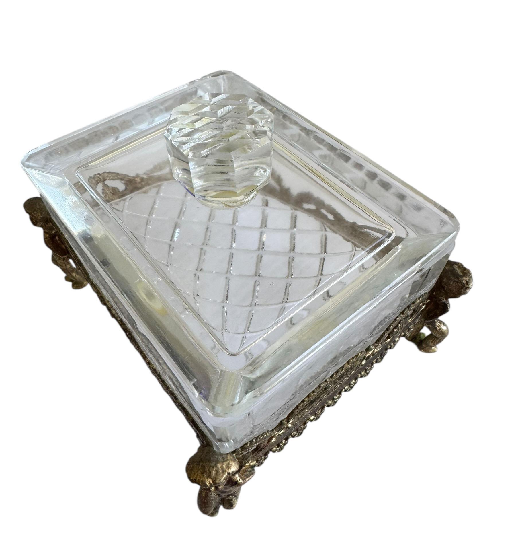 An exquisite French crystal and bronze dresser box featuring a detached lid topped with a large, cut crystal finial. The box is supported by four charming bronze cherubs. Beautiful bronze swags drape around the crystal body, enhancing its luxurious
