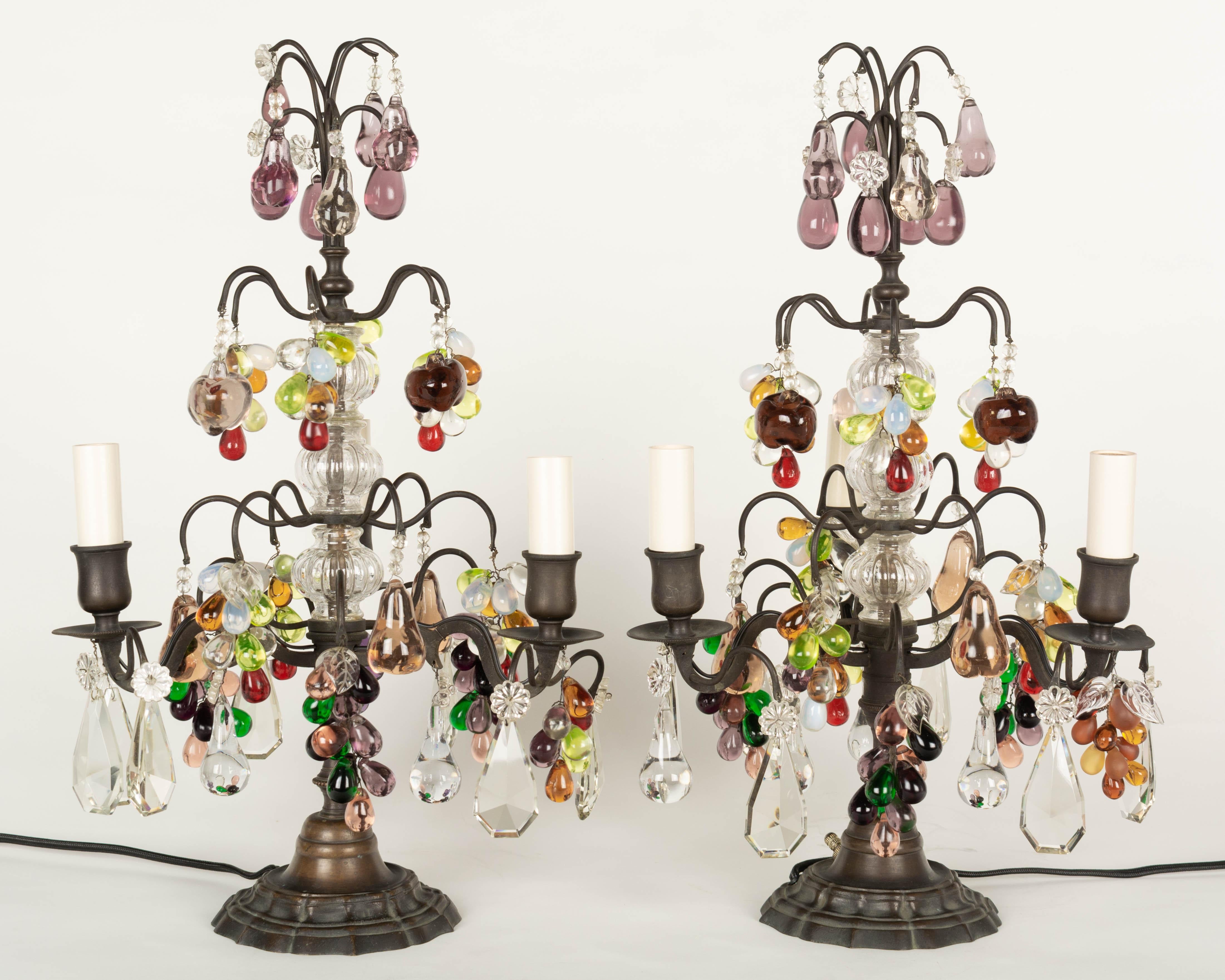 A pair of Louis XV style three-light French girandoles, or chandelier table lamps. Dark antique brass frame with three tiers of crystal prisms with rosettes. Colorful green, opal, amber and amethyst glass grape clusters, pears and apples. Rewired