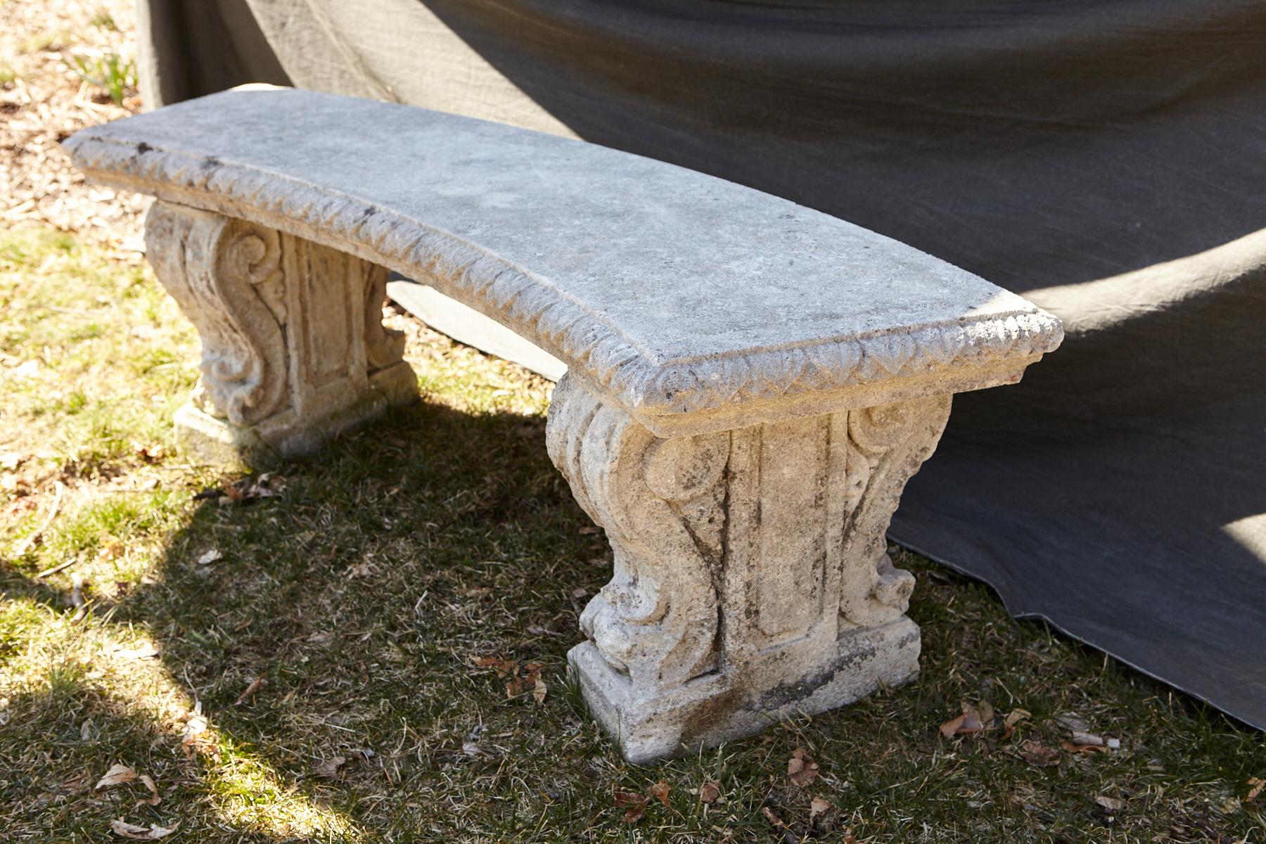 Curved cast stone bench with an ornamental carved band of alternating concave and convex sections around the flat seat, reminiscent of flower petals, mounted on carved legs with carved double sided scrolled acanthus leaves.
The bench has a very nice
