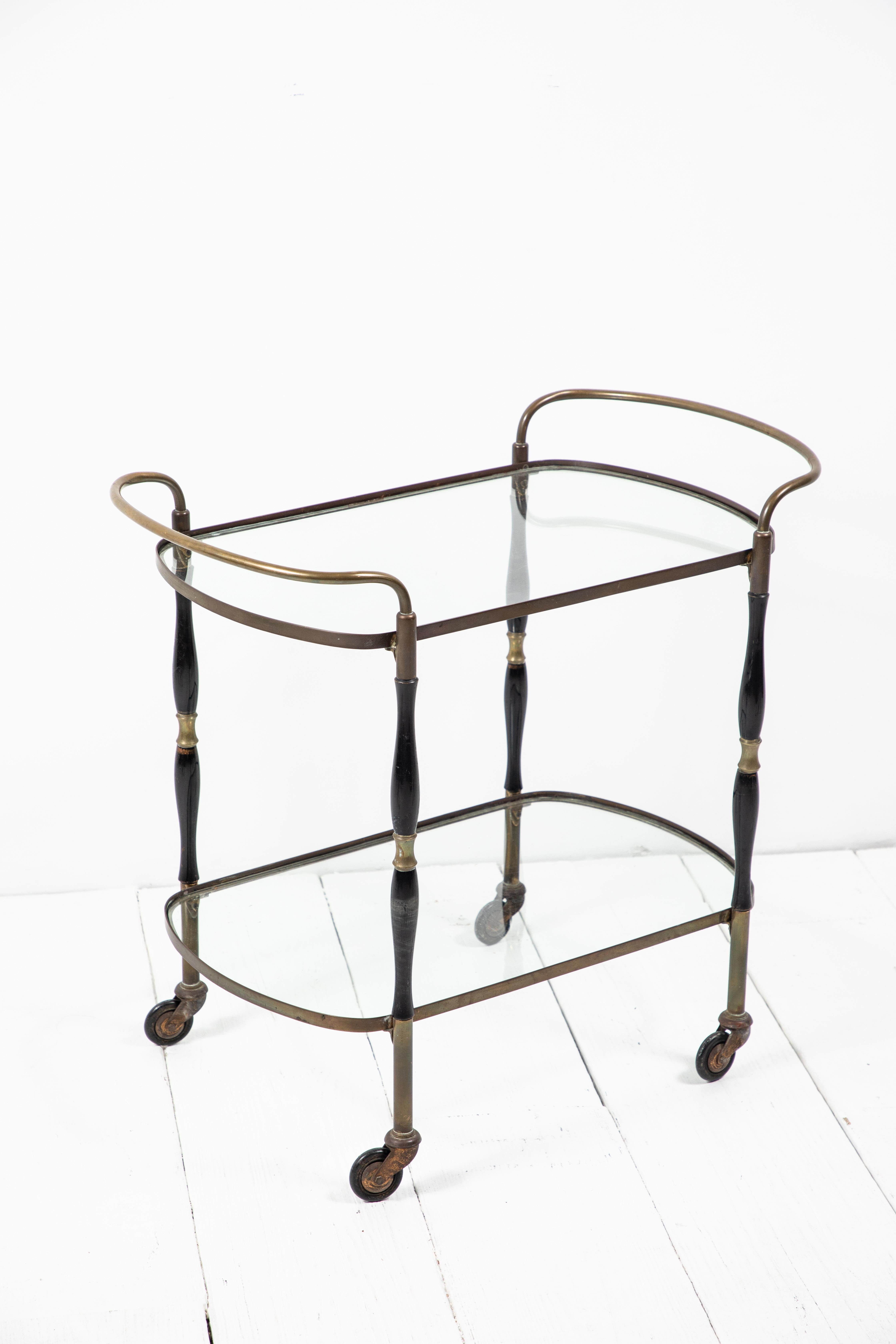 Beautiful deco bar cart with black and brass turned legs with two glass shelves. The bar cart sits on wheels.