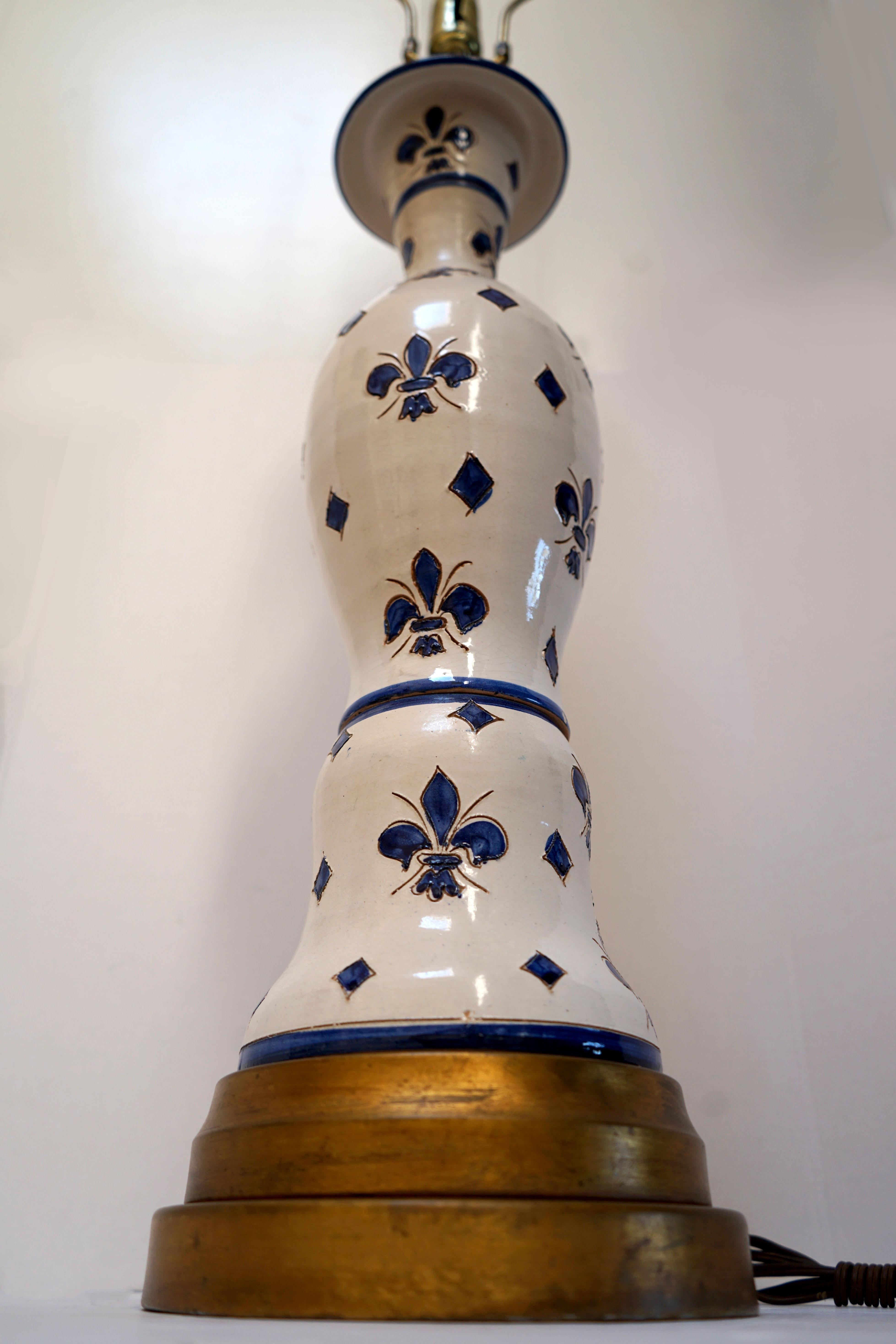 Blue and white baluster shape and delicately carved fleur de lys in blue on white ceramic is reason enough to covet this beautiful oversized table lamp produced between 1940 and 1960. The collar like neck complements the gold circular wooden base.