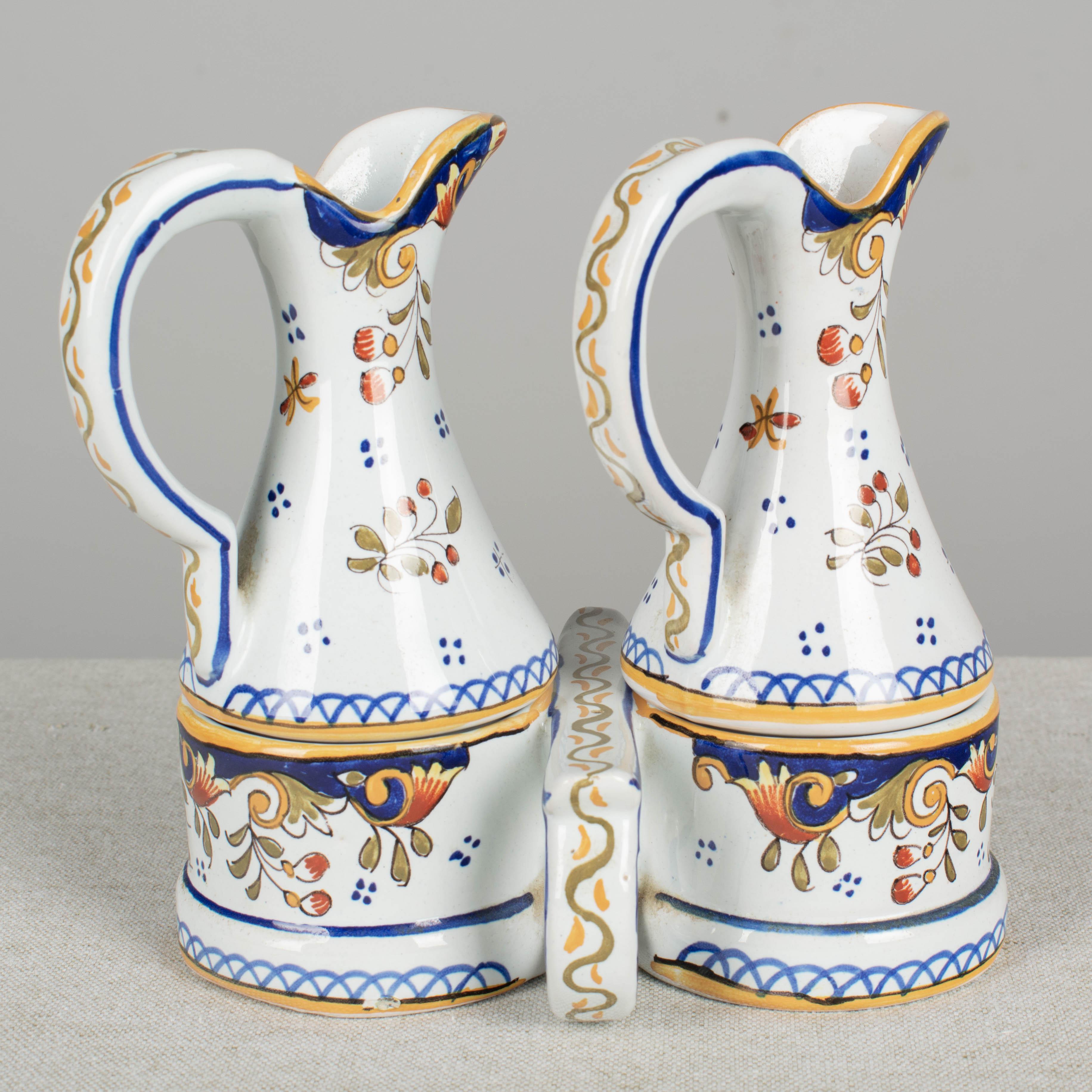 A French Desvres faience cruet set, with hand painted floral decoration in the traditional colors of blue yellow, green and orange. The base holds a pair of handled pitchers, for oil and vinegar, each decorated with the crest of Normandy, a red