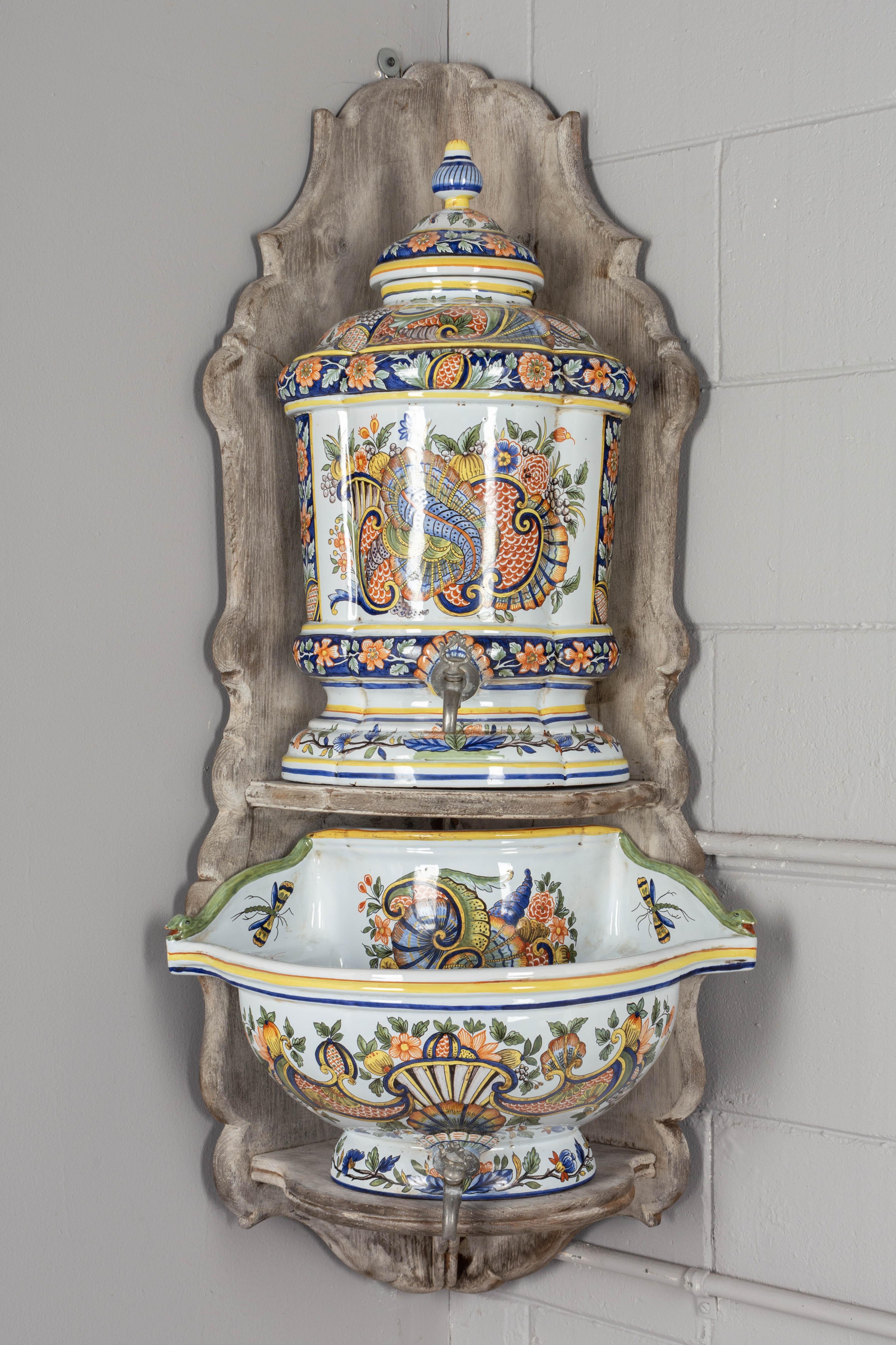 A French Desvres faience Louis XV style lavabo with traditional colorful hand painted decoration. The lidded urn and large deep basin rest on a whitewashed chestnut wall mounted corner shelf. Each with metal spouts and makers mark on the underside.