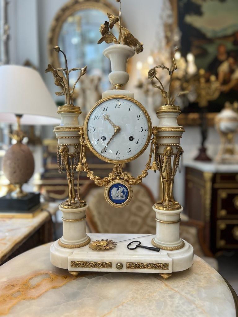 Directoire portico clock decorated with two Athenian with eagle heads and feet. 
The three gilded bronze hats are decorated with flowers. The white Carrara marble Medicis vase sits above the dial. 
The pendulum is decorated with a sun. The Athenian