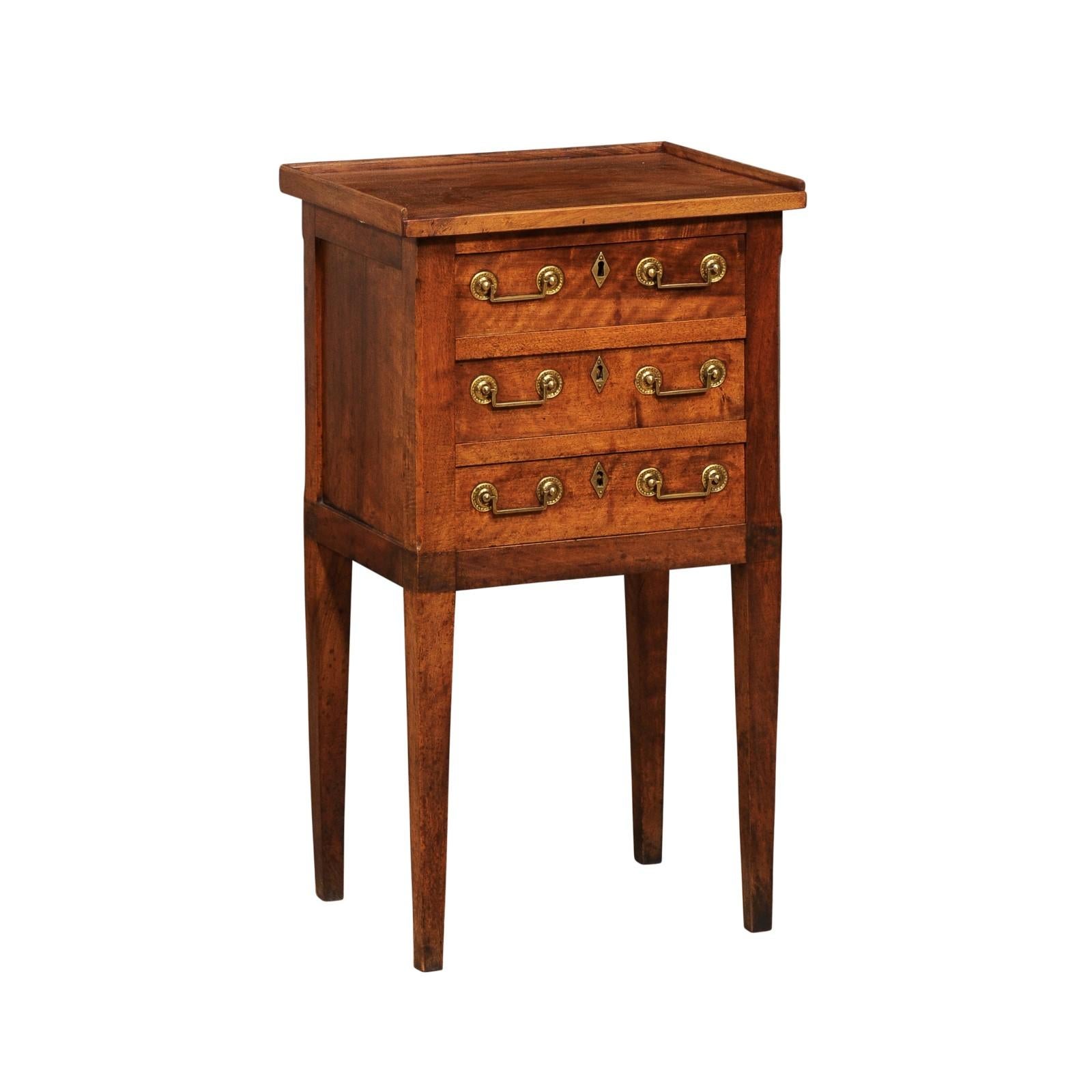 A French Directoire period walnut bedside table from the 18th century with three drawers, tapered legs and brass hardware. Steeped in the elegance of French Directoire design, this 
