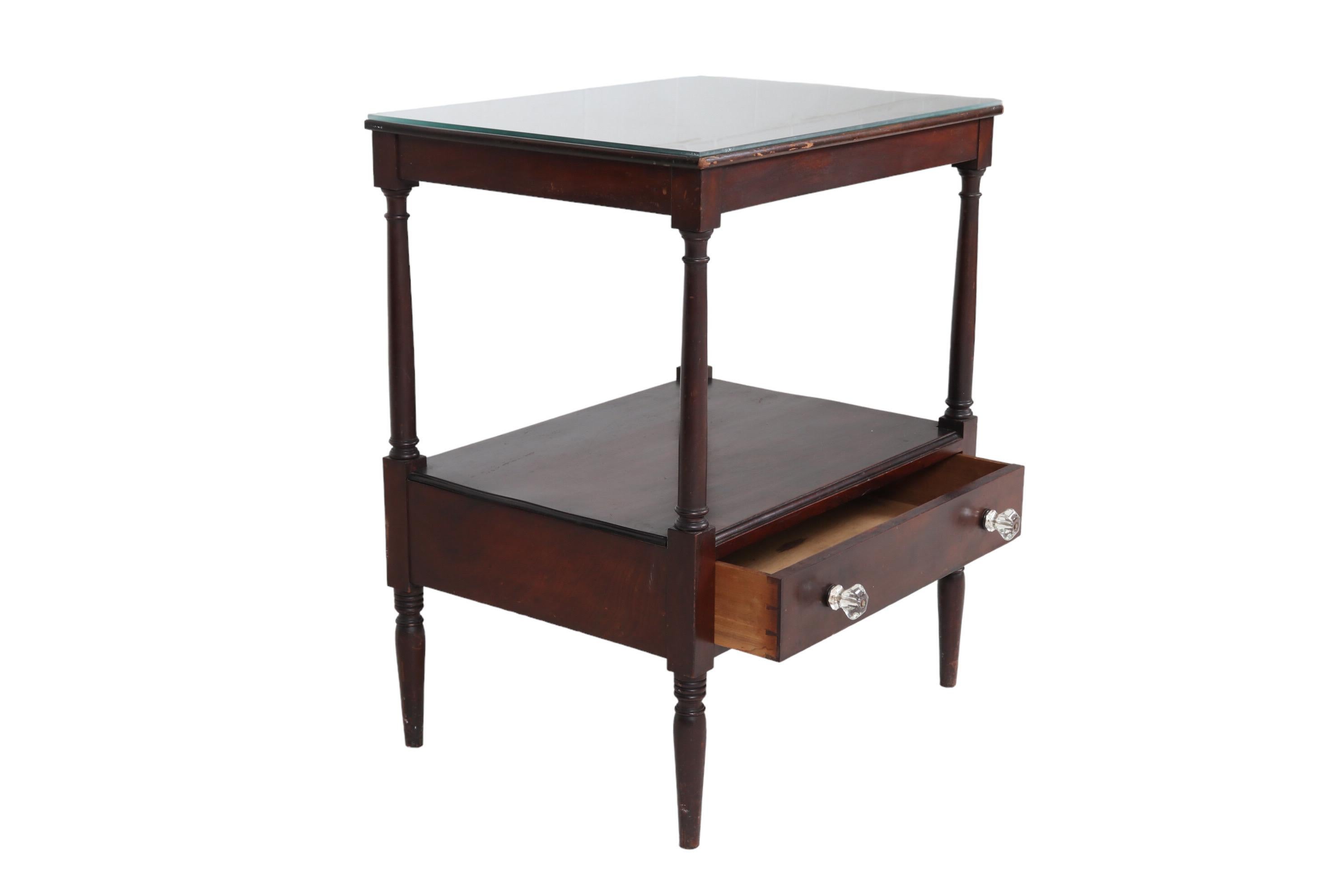 A French Directoire period side table made of mahogany. The tabletop is finished with a beveled edge and has a protective glass cover. Below, a single drawer is hand dovetailed and opens with two round glass knob handles. Supported above and below