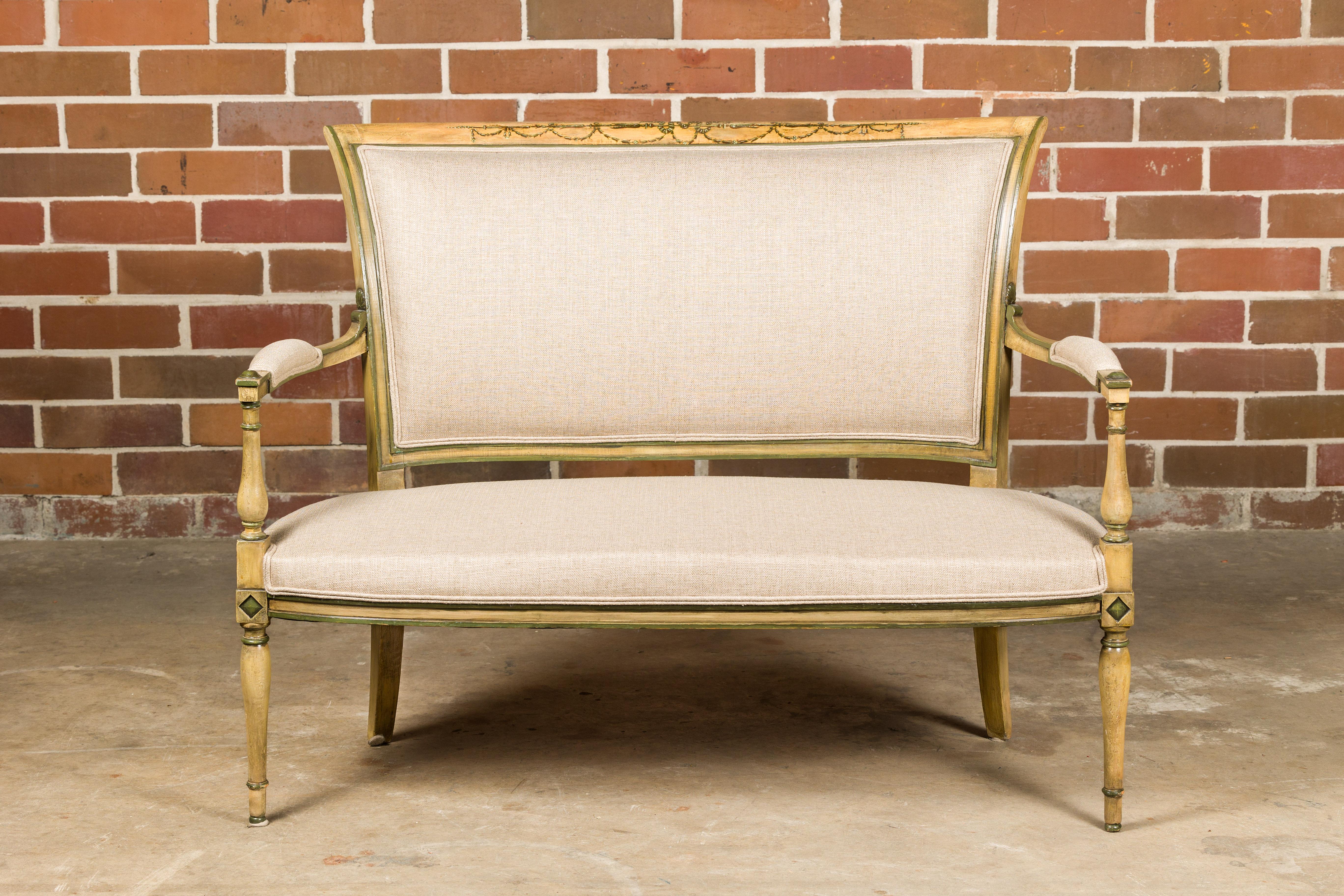 A French Directoire settee from the 19th century with soft creamish yellow painted finish, green accents, painted garlands and linen upholstery. Immerse yourself in the graceful charm of this 19th-century French Directoire settee, a masterpiece that