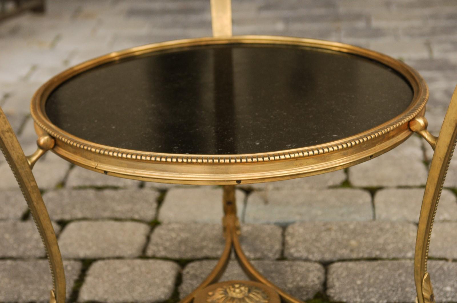 A French Directoire style gilt bronze guéridon table from the early 20th century, with black marble top and lower shelf, ram's heads, hoofed feet and beaded motifs. Born in France during the Roaring Twenties, this superb guéridon table features a