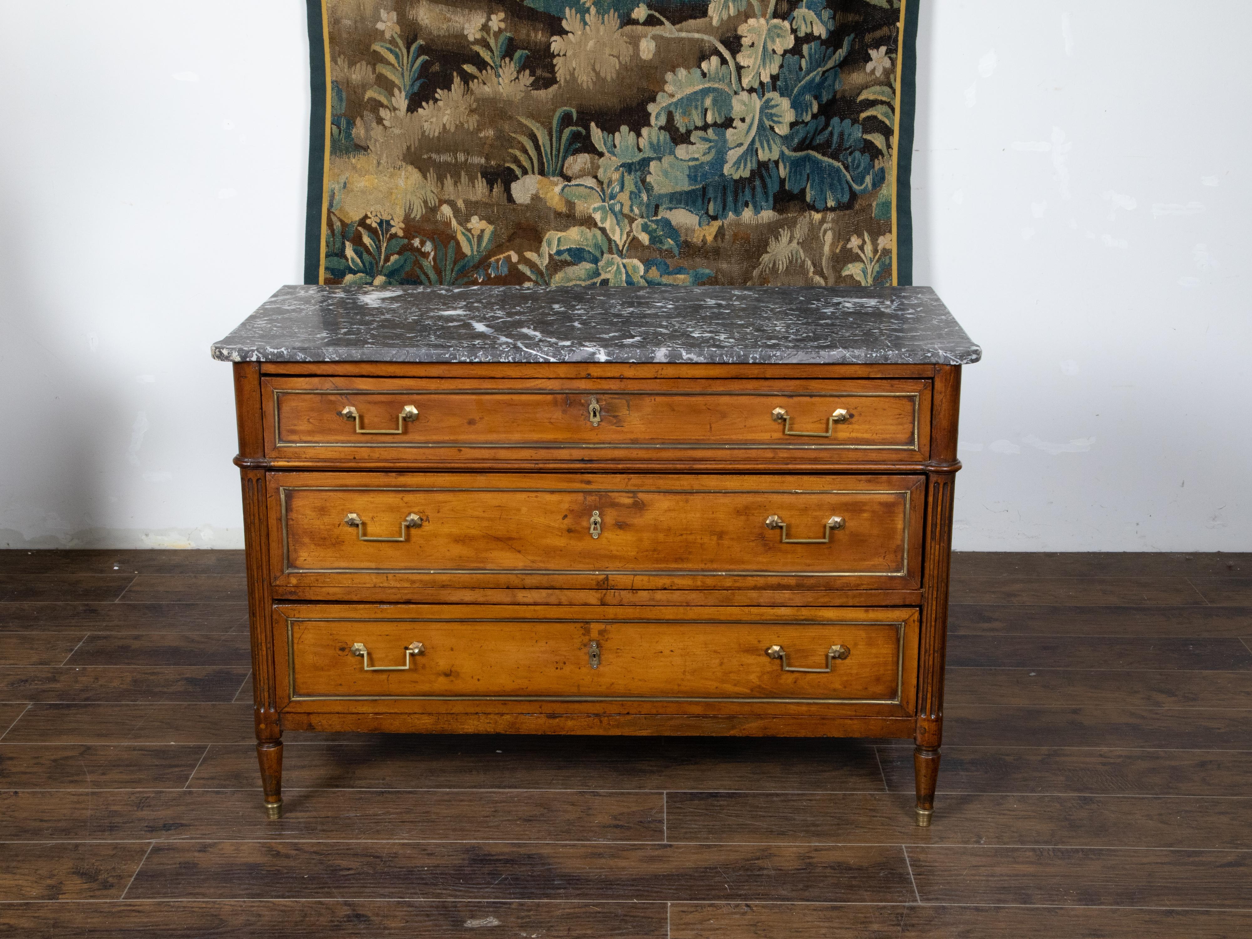 A French Directoire style walnut commode from the 19th century, with three drawers, grey marble top, brass accents and fluted posts. Created in France during the 19th century, this walnut commode showcases the stylistic characteristics of the