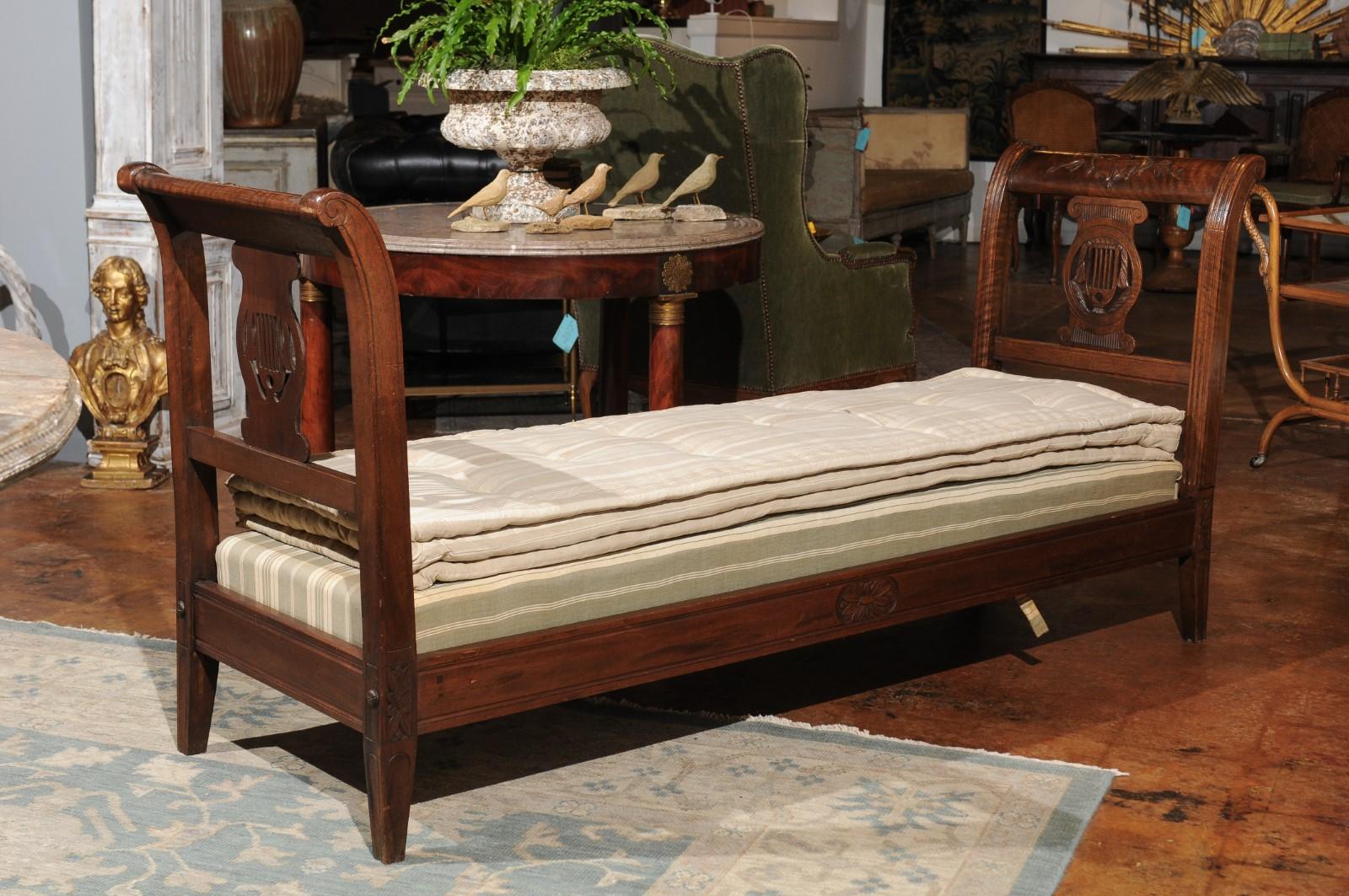 A French Directoire style walnut daybed from the 19th century, with carved lyre and urn motifs. This exquisite French Directoire style daybed features an un-upholstered rectangular seat with cushion, flanked by two elegant out-scrolling arms. Each