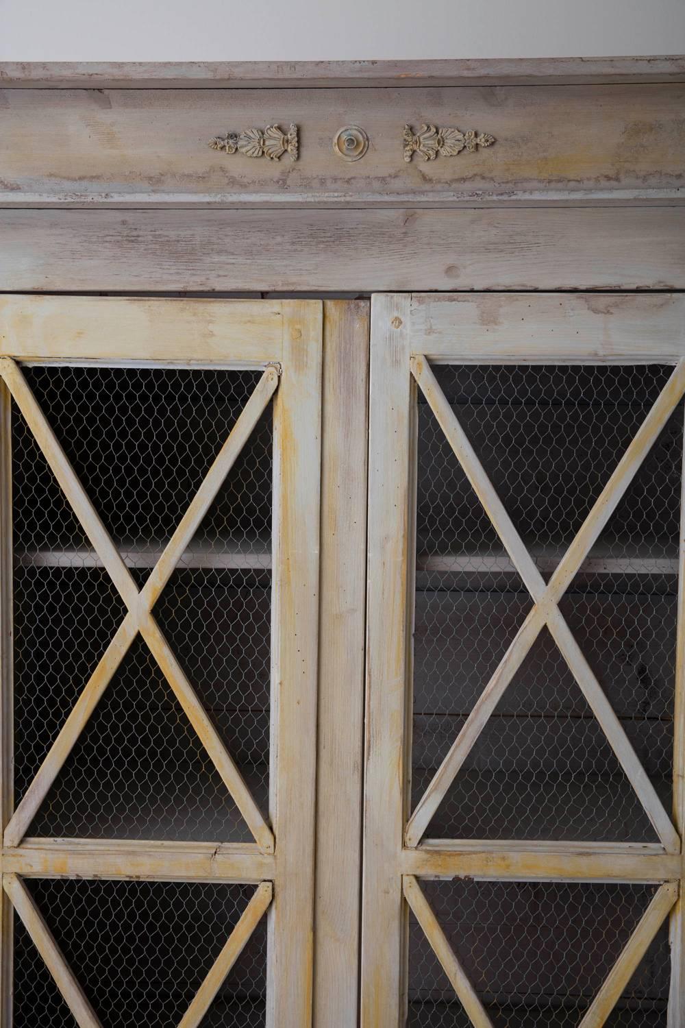 French bookcase or armoire in the directoire style, painted in shades of grey with a wash of soft yellow, chartreuse, and a charcoal grey painted base. There is a corniced top over two full-length doors fitted with chicken wire with four removable