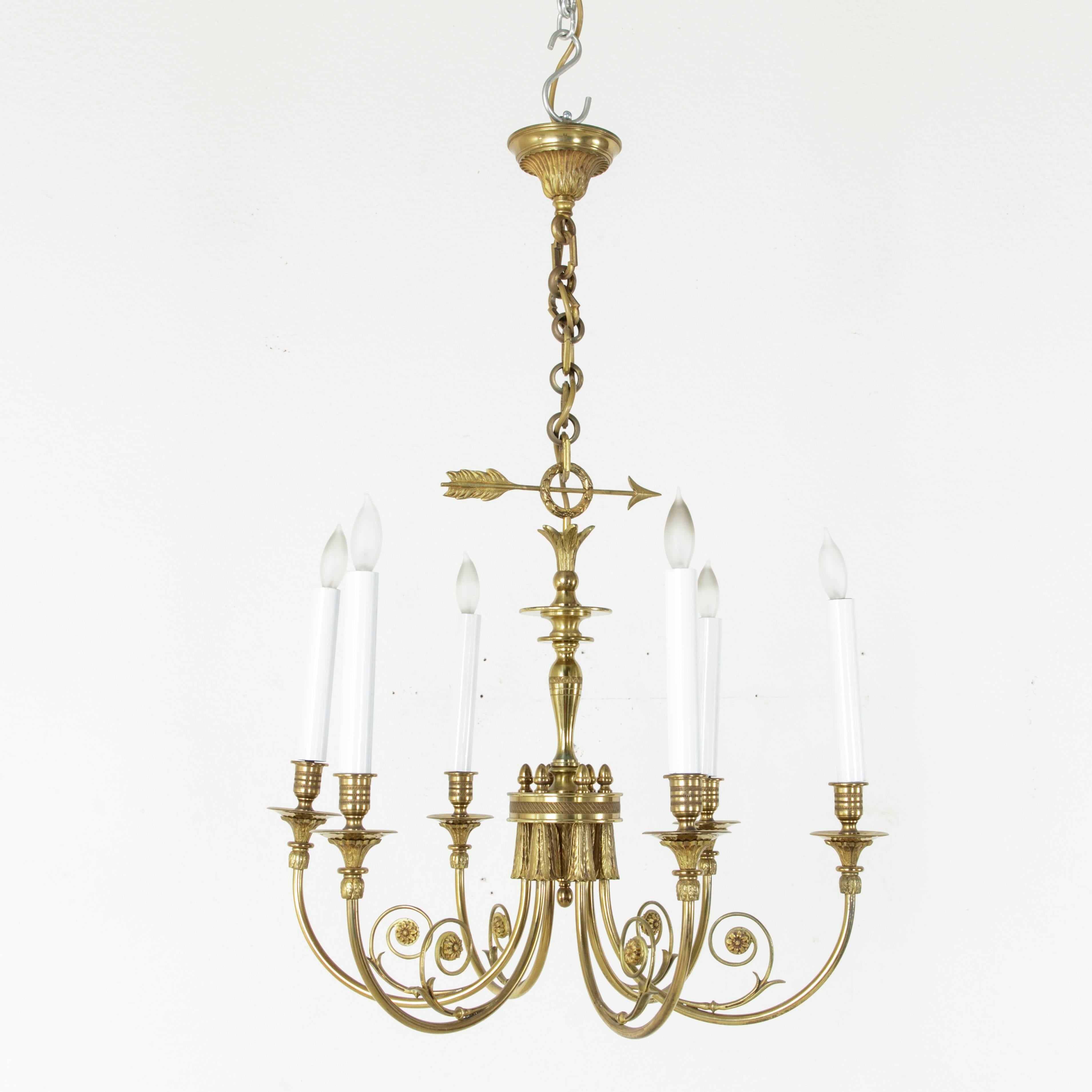 This elegant and classic bronze Directoire style French chandelier is of brightly polished solid bronze. The six arms each feature a curling frond ending in a single rosette while the central column is detailed with feather and arrow finials. Each