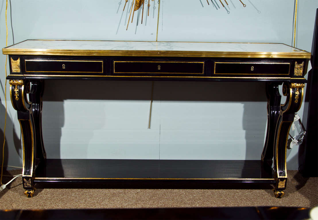 Glamorous French Directoire style ebonized and parcel-gilt console table, circa 1940, the white marble with bronze banding over a narrow frieze fitted with three drawers, over scroll-decorated legs joint by a rectangular lower tier, raised on gilded