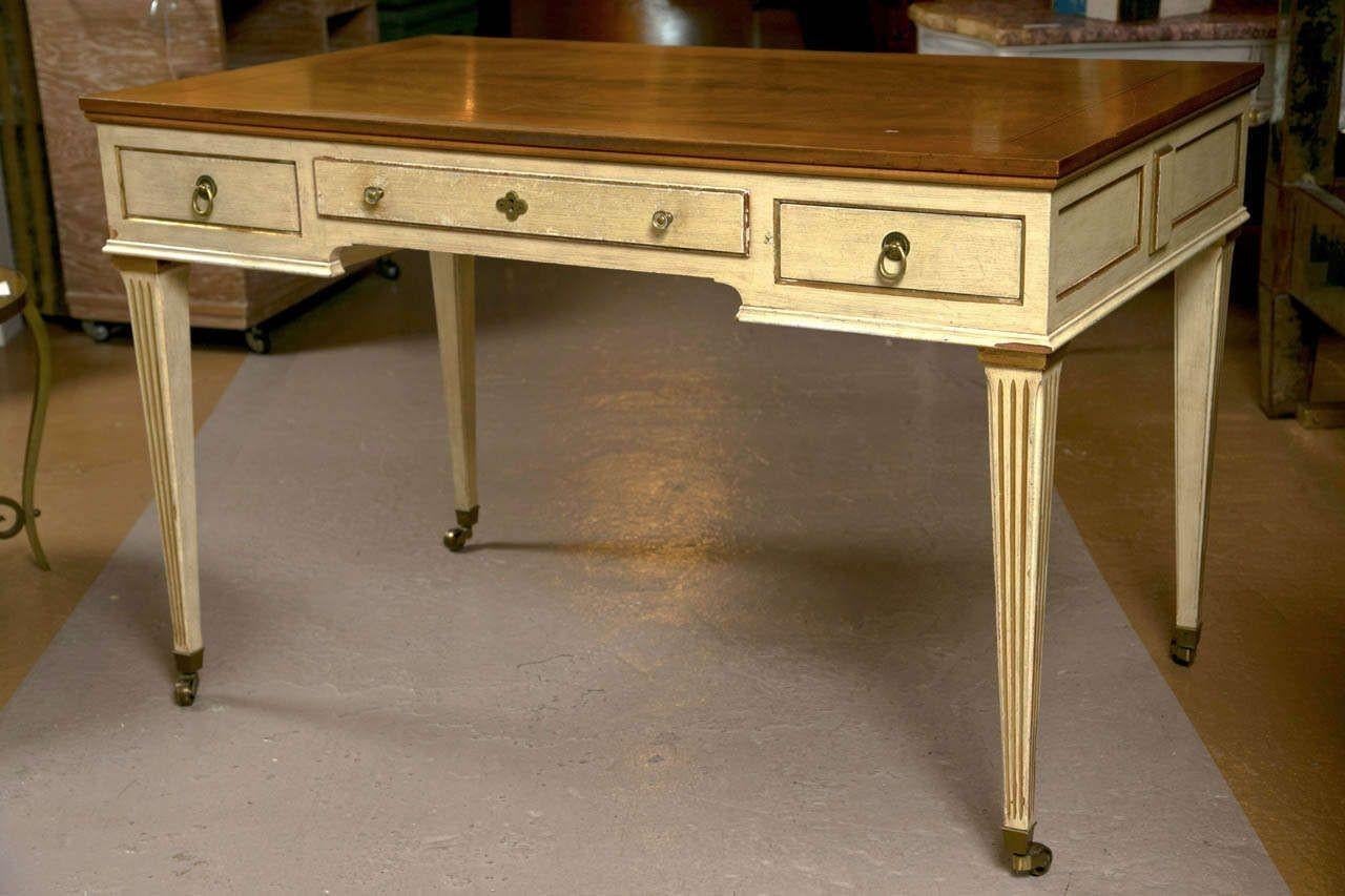 A creme peinte bureau plat in the style of French Directoire, circa 1960s, the wood top in natural finish over a frieze fitted with three drawers and decorated with parcel-gilt banding, supported on square-tapering fluted legs ending in