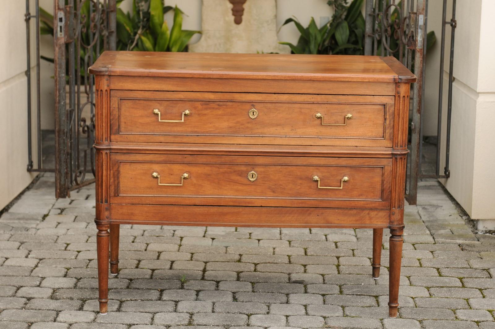 A French Directoire style walnut veneered two-drawer commode from the first half of the 19th century with brass hardware, fluted side posts and turned legs. This French walnut commode features a rectangular top with rounded corners in the front,