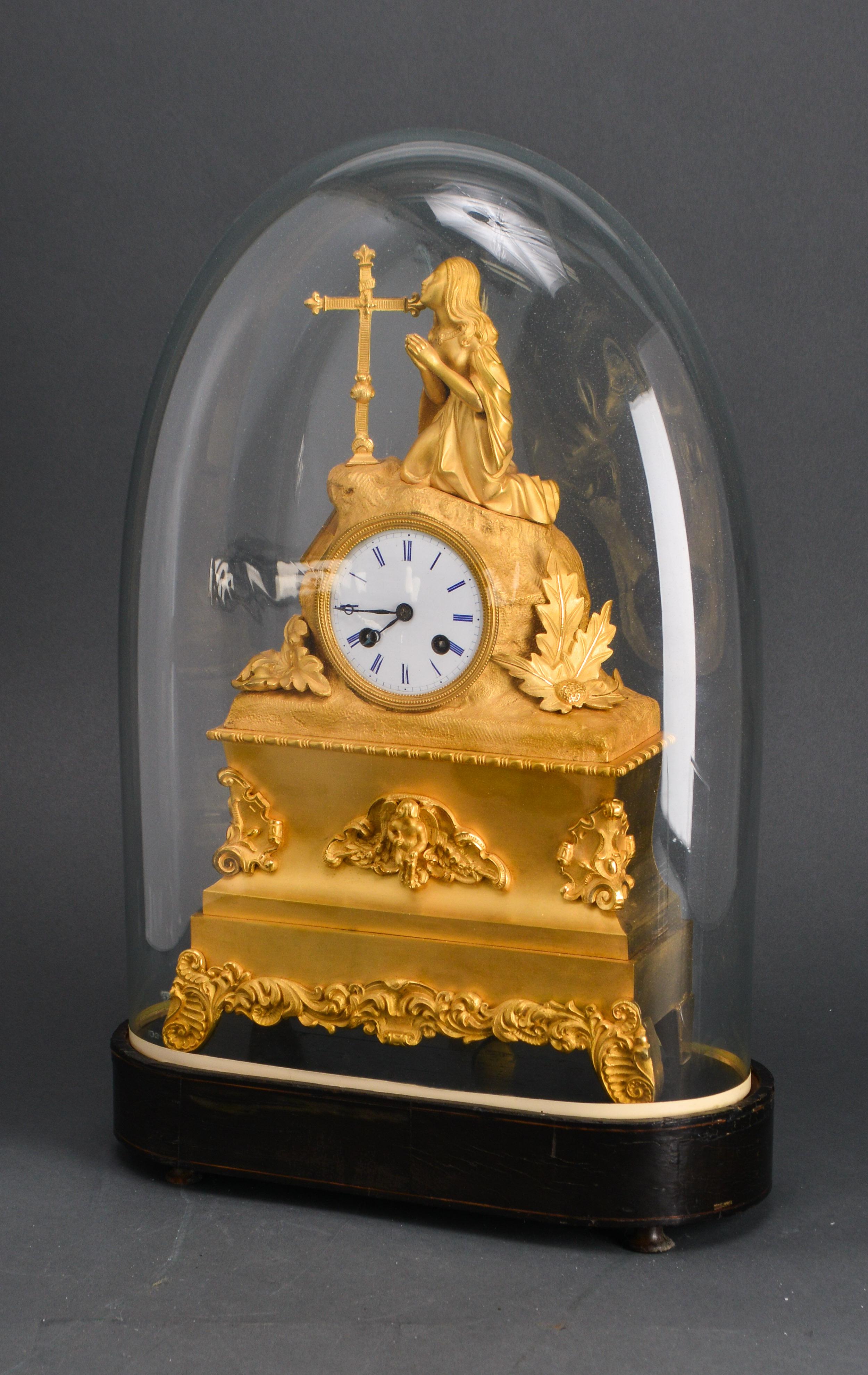 French gilt ormolu bronze chiming clock with white porcelain dial with black hands and blue Roman numerals and a kneeling praying female figure in front of a cross. The piece is housed under a Victorian glass dome and dates from the late 19th