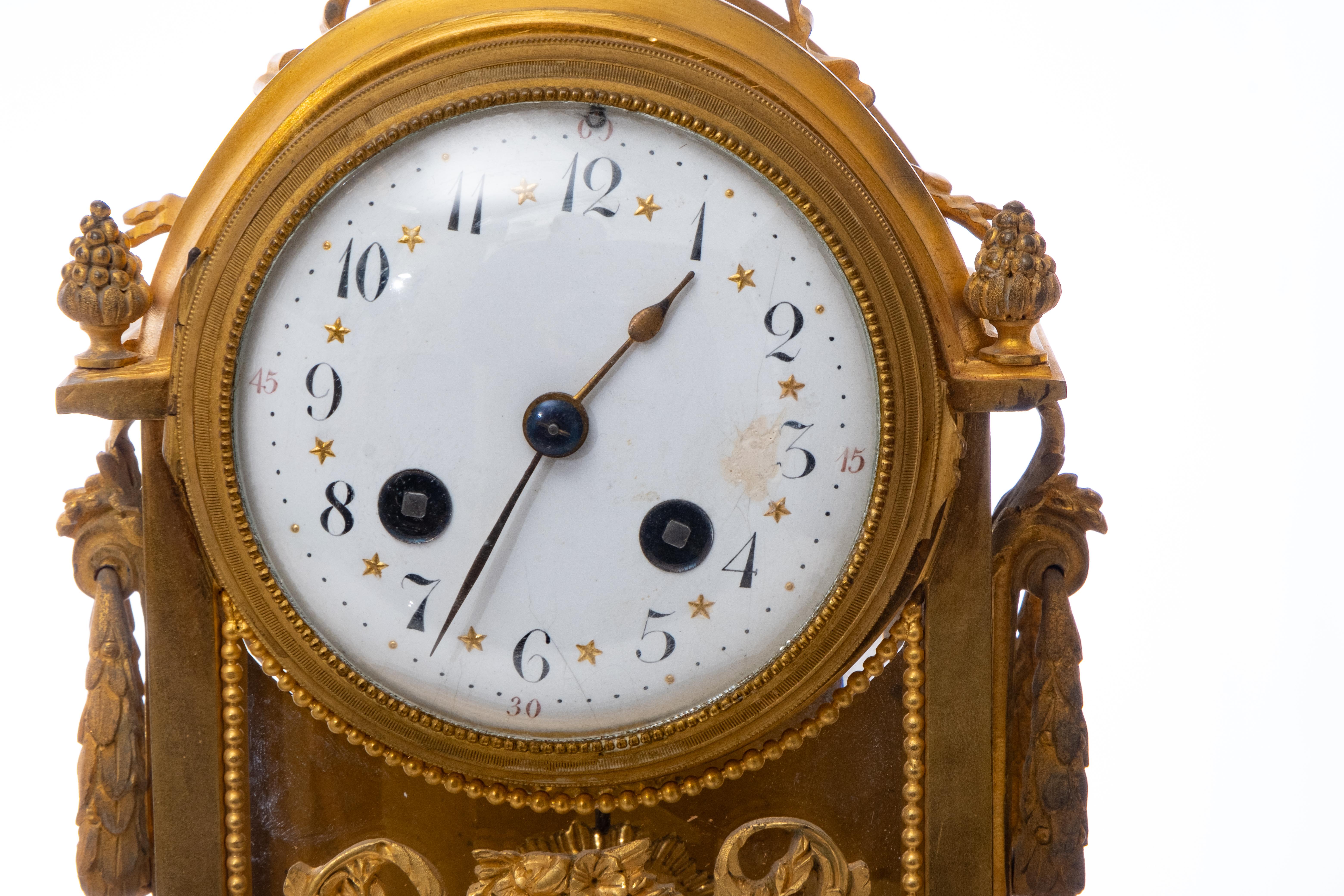 An antique 19th century French dore bronze clock and candlestick garnitures set. Footed clock features glass panel sides and back, foliate ormolu garland, and flower wreath motif, topped with crossed troche in a wreath finial. White enamel clock