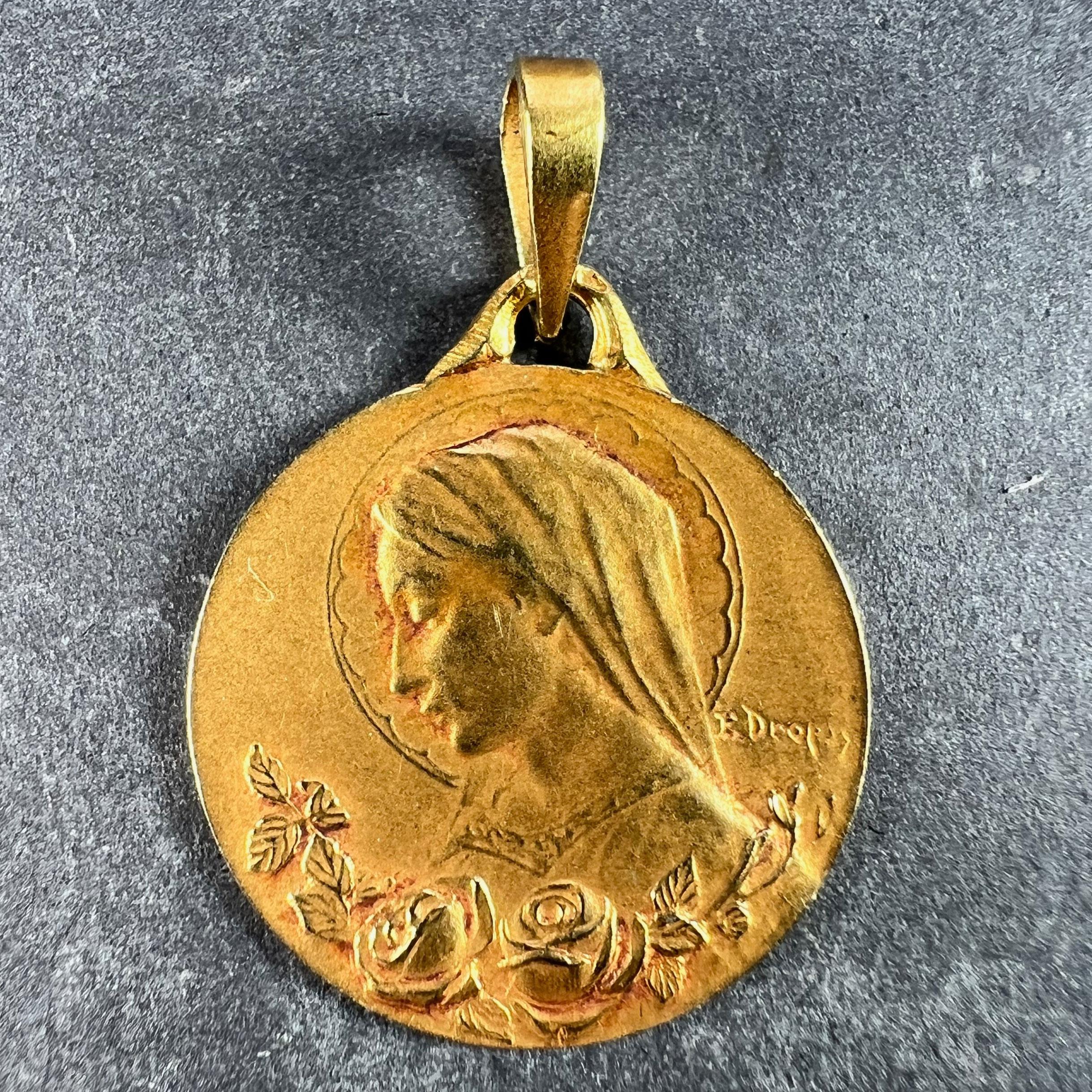 A French 18 karat (18K) yellow gold charm pendant designed as a medal depicting the Virgin Mary in profile with a halo above a pair of roses in full bloom. Signed E. Dropsy. The reverse shows a sheaf of lilies, a symbol of purity. Stamped with the