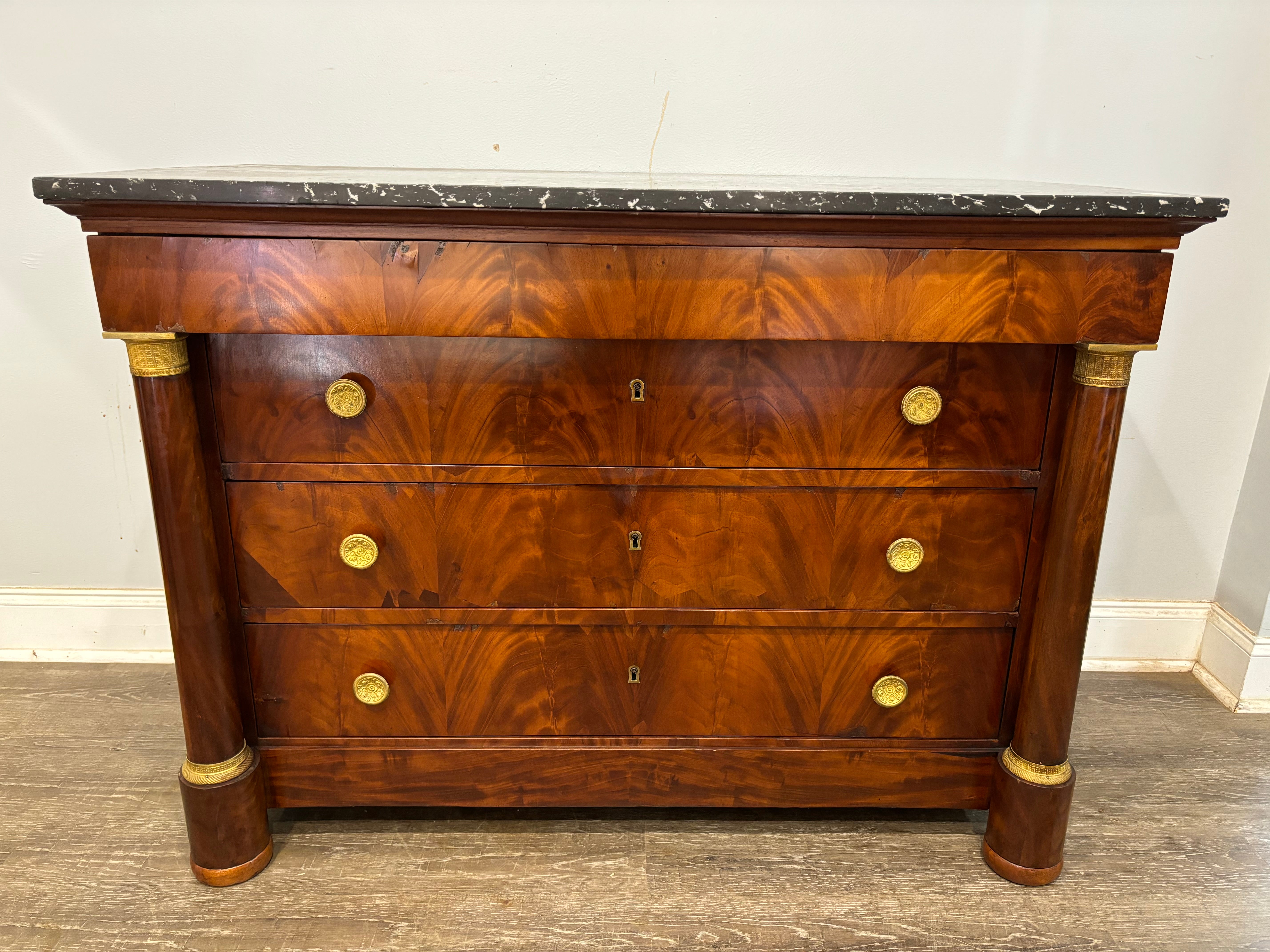 Cette commode Empire française est en acajou flammé avec un plateau en marbre d'origine. Il possède ses ferrures et ses anneaux d'origine autour des colonnes.
