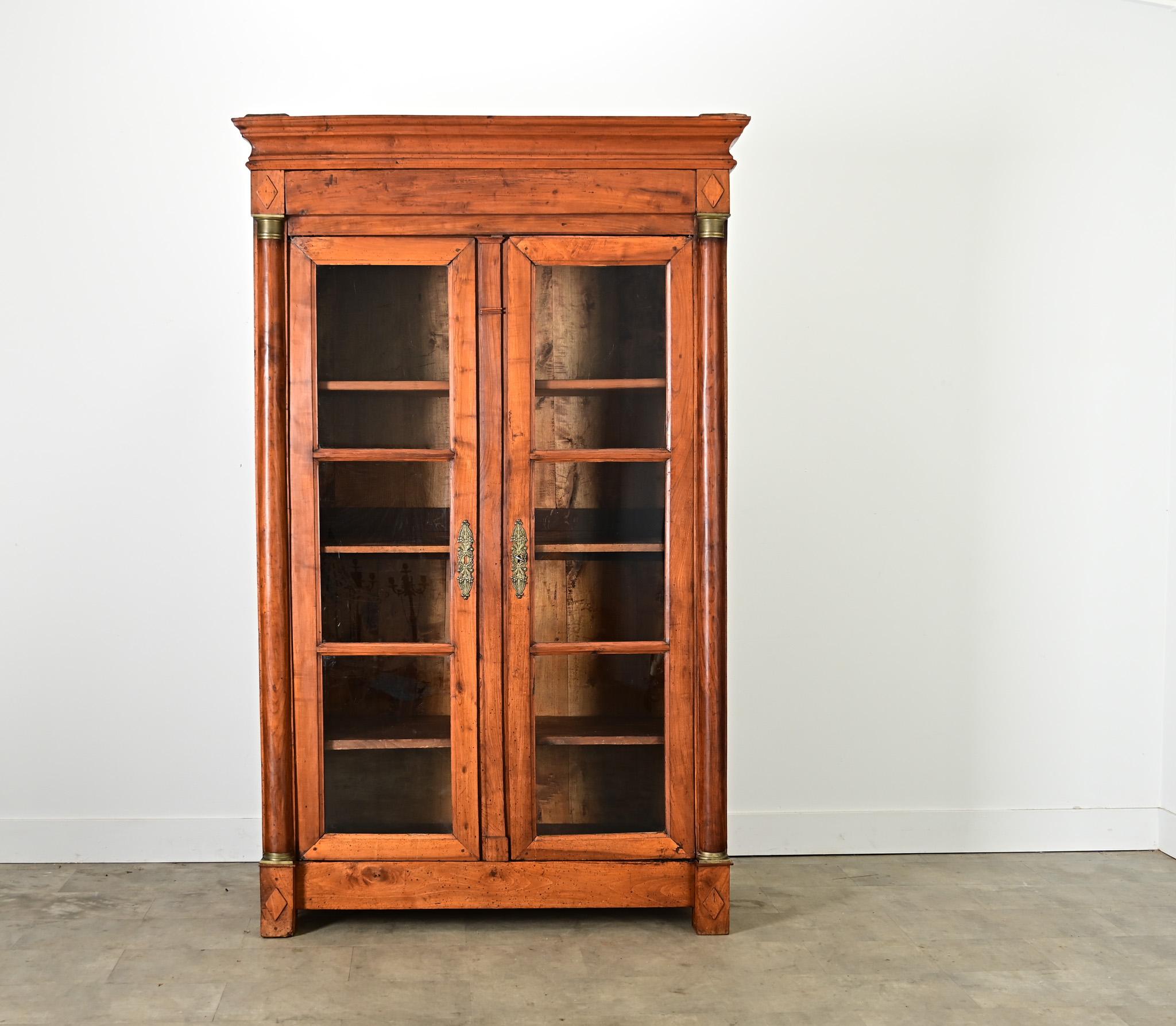 A 19th century French Empire style bibliothèque or display cabinet, handcrafted using solid fruitwood and two glass-front doors accented with brass. The clean, straight lines allow this handsome design to fit any number of traditional and