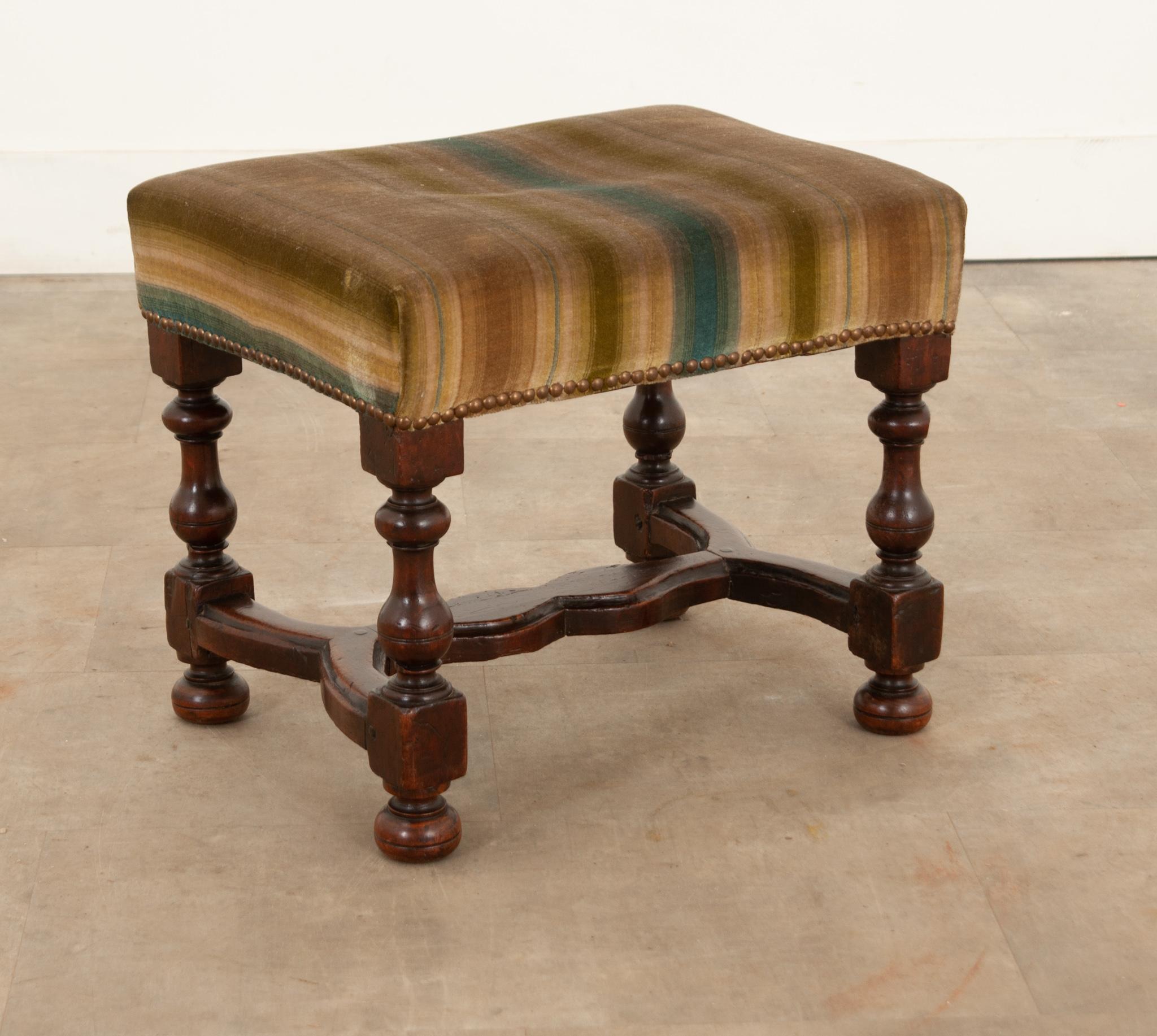 This stately yet petite stool is the perfect antique to add interest to any space. Upholstered with brown, olive, and teal striped velvet  in excellent, used condition and held with round brass rivets. The heavy solid walnut frame showcases