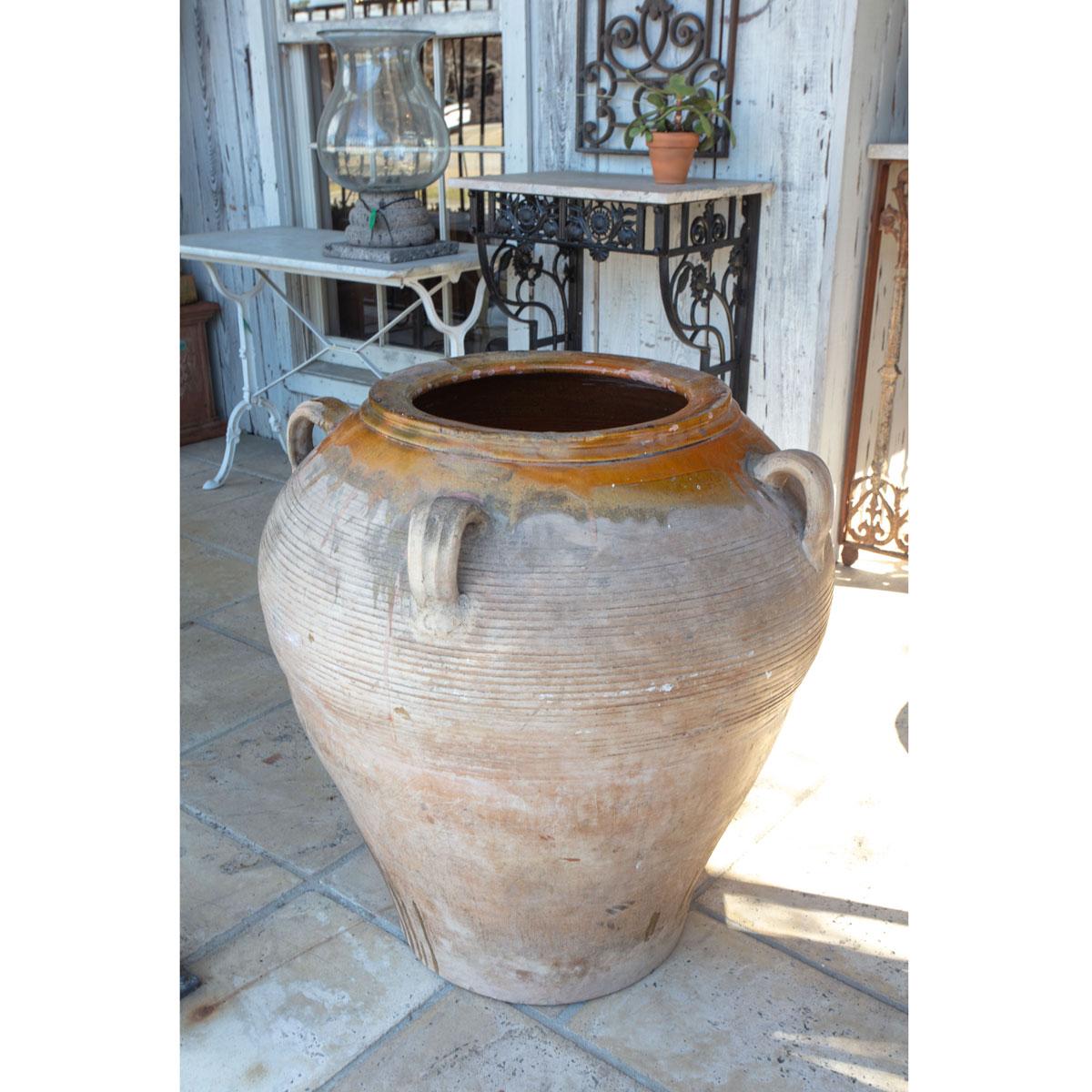 This large clay olive jar has been partially glazed in an earthen orange/yellow color. There are four handles equally spaced around the top of the jar and a ribbed pattern wraps the jar just below the handles. Make a statement on your porch or in