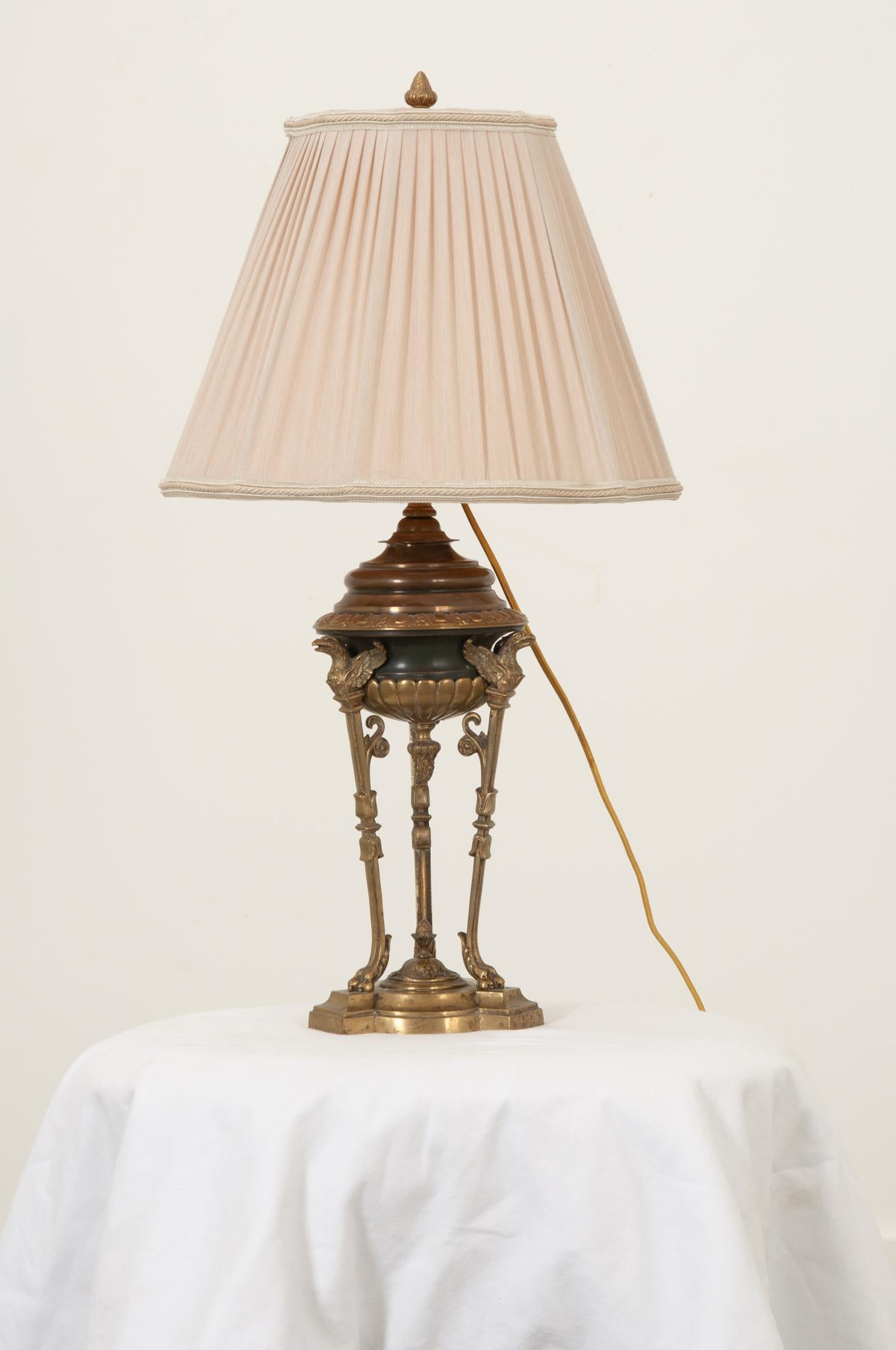 An early 20th French Empire style table lamp has a concave tri-form bronze plinth base set atop a slightly larger piece of marble in the same shape. The upper bowl portion is supported by three brass legs with paw feet topped by three brass gryffon