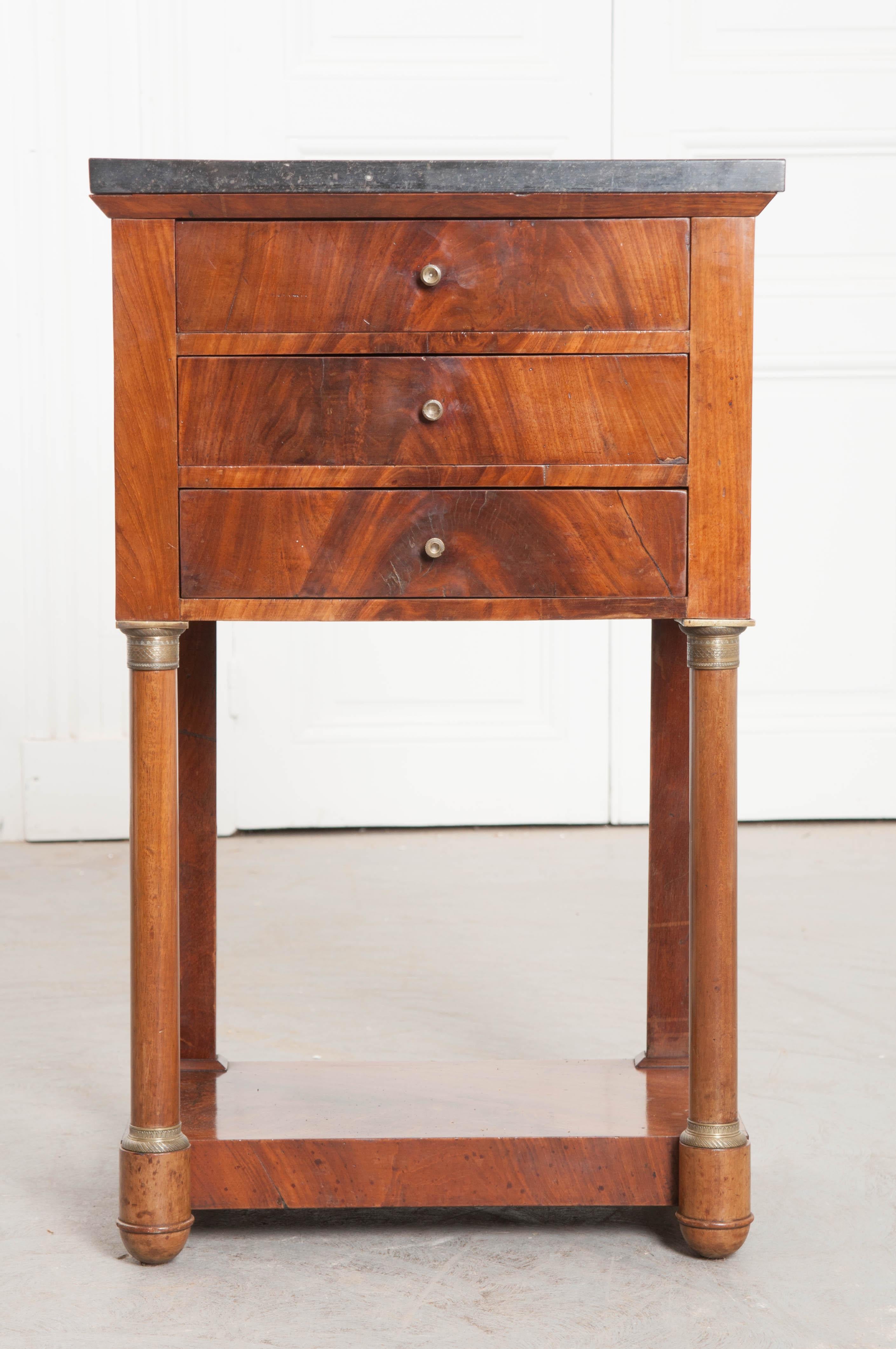 A stately three drawer mahogany bedside table, with black fossil marble top, from 1910s France. The table was made in the Empire style using beautifully toned mahogany with exceptional grain. The rectangular piece of black fossil marble is in