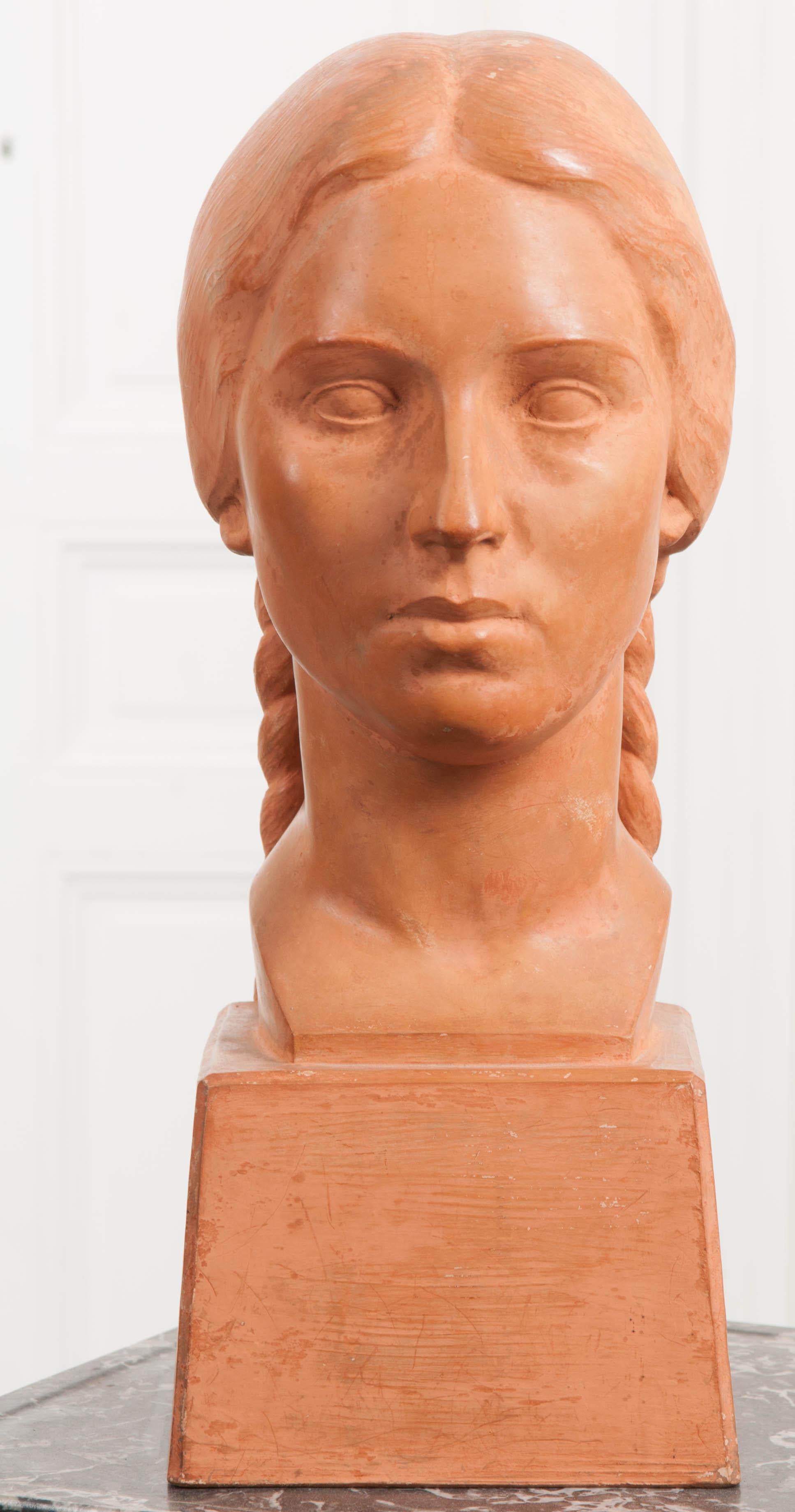 A distinguished terracotta bust from the early part of the 20th century. Made in France, the work depicts a female with long, braided pigtails and her hair parted down the center. An unemotional expression is worn on her face, now frozen in time.