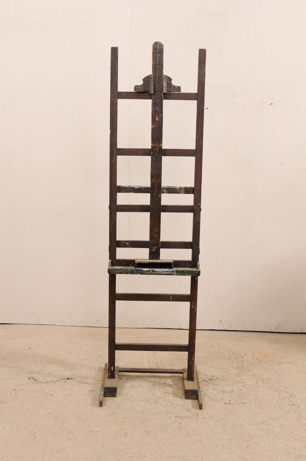 A French wooden adjustable artist easel from the early 20th century. This good-sized antique easel has a small supply shelf, support for a painting, and an adjusted frame. This piece is supported by a H-shaped braced feet. This rustic French easel