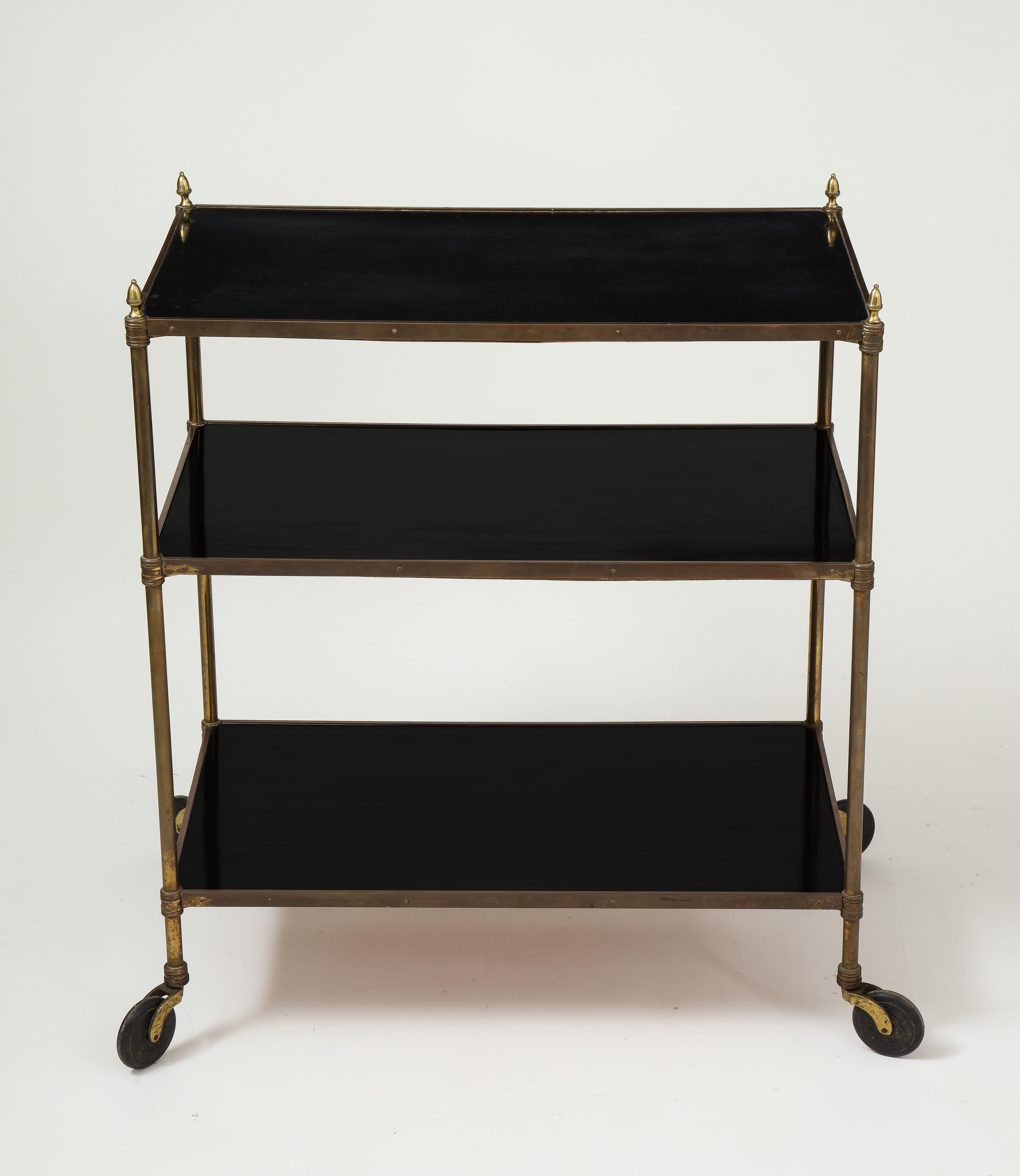 With three rectangular ebonized wood shelves set within gold-lacquered brass supports, the corners surmounted by cone-shaped finials; on wheels.