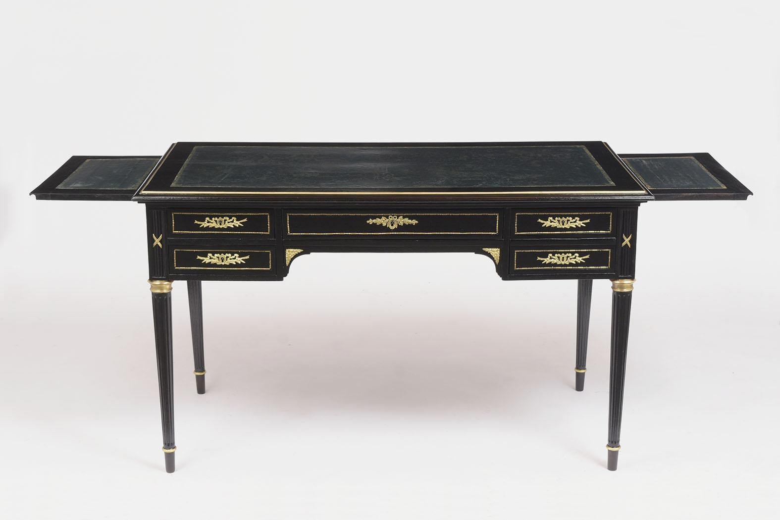This 1880s Antique French Louis XVI-style desk is made of mahogany wood with a deep ebonized and lacquered finish. The top features the original embossed leather insert with gilt details. There are five drawers with brass molding and large key