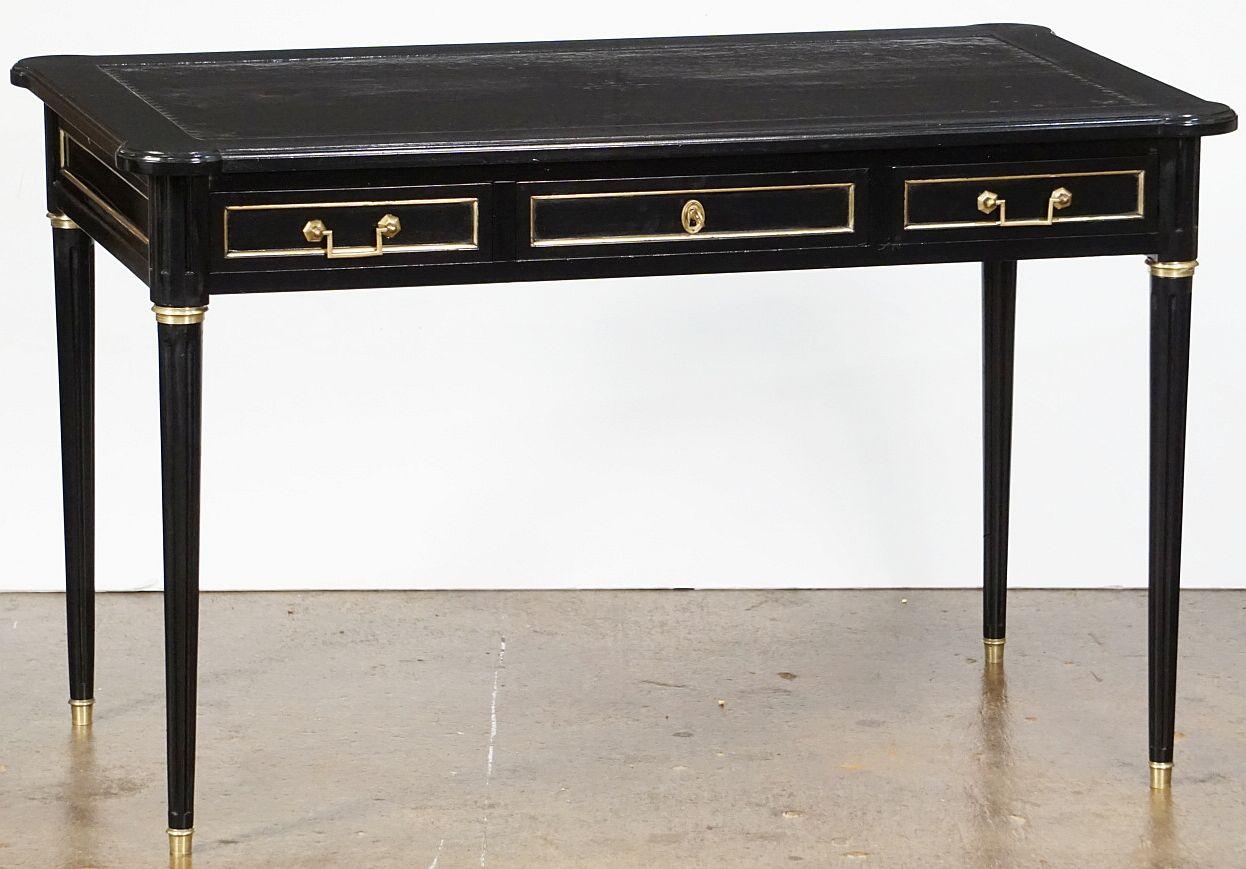An elegant French bureau writing desk (or table) of ebonized mahogany, in the Louis XVI style, featuring an embossed leather top with gilt bronze accents over a frieze with fitted drawers, with two pull-out writing leaves to right and left. Resting