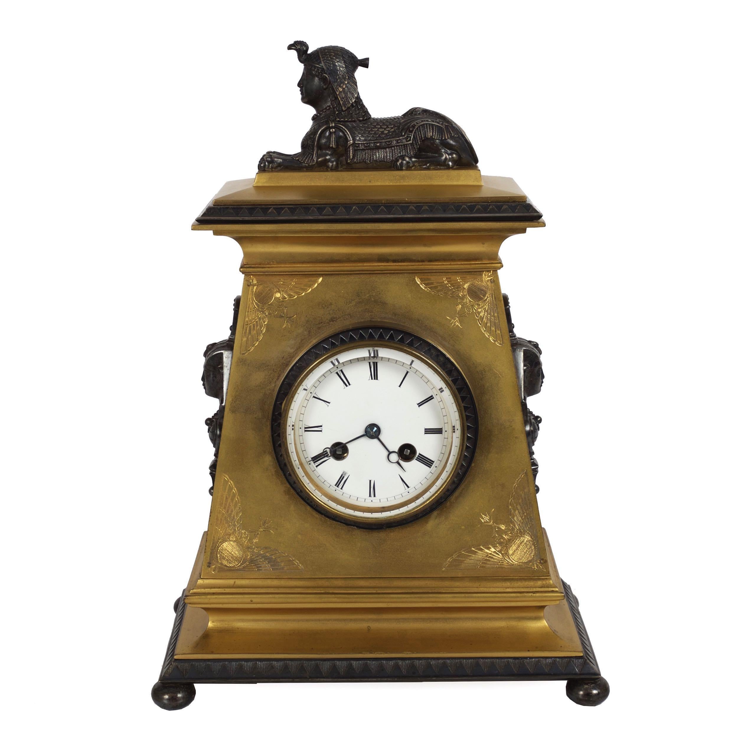 A superb Neo-Egyptian mantel clock characterized by a trapezoidal gilt bronze body with spandrels incised with twin phoenix motifs surmounted by a silver plated bronze seated sphinx and flanked on either side by silver plated Pharaoh masks. The