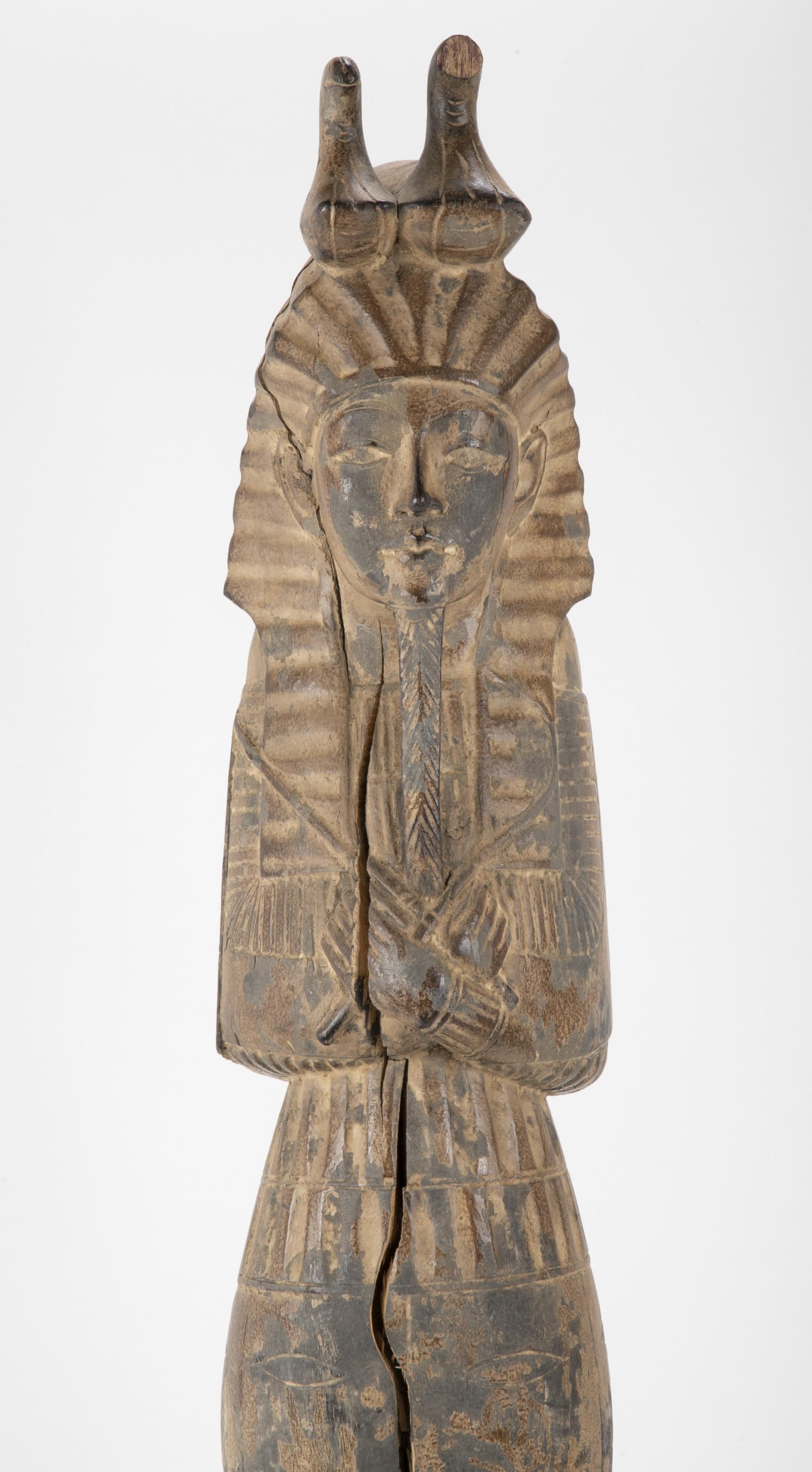 A carved standing figure of King Tutankamun wearing the royal headdress topped by the double uraeus. This compelling carving depicts the slender boy king standing with arms crossed holding a crook and a flail. The worn surface of the piece and the