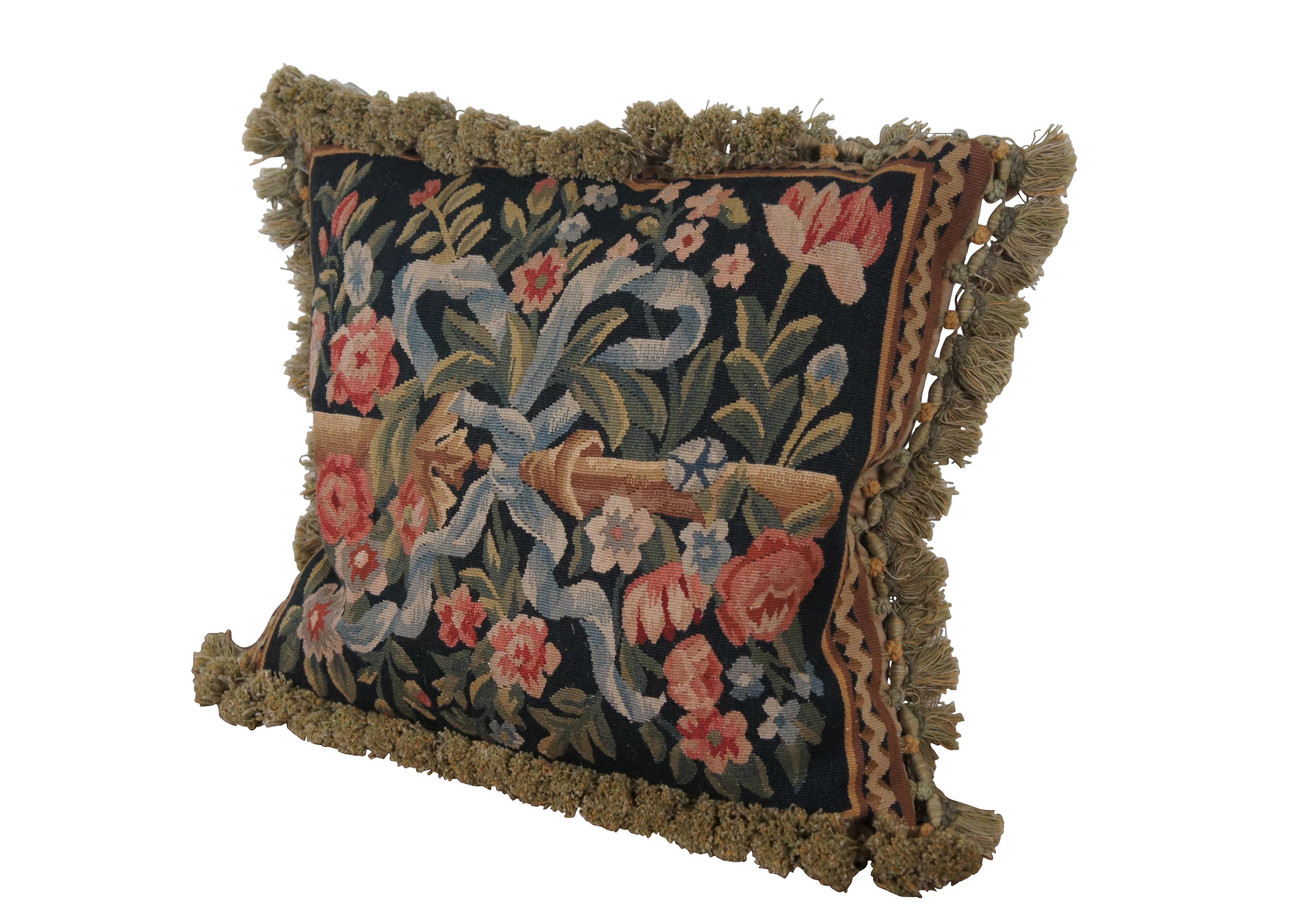 20th century square throw pillow, embroidered with blue flowers and pink roses on leafy stems, surrounding a golden bar tied with a blue ribbon / bow, all on a deep navy blue background. Olive green and gold tassel trim. Brown velour back with