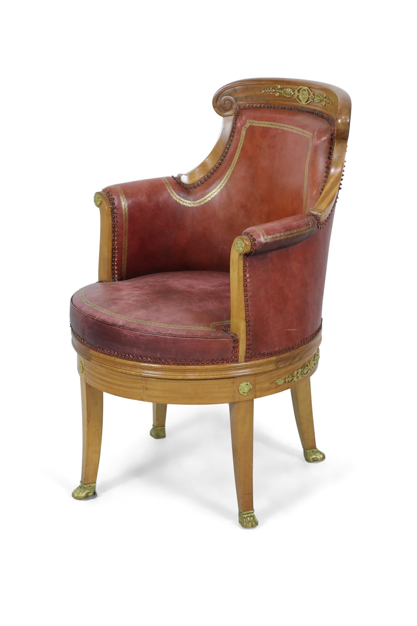 French Empire blond mahogany swivel armchair upholstered in gilt embossed red leather with brass nail head trim and having bronze embellishments and medallions throughout with hoof sabots.