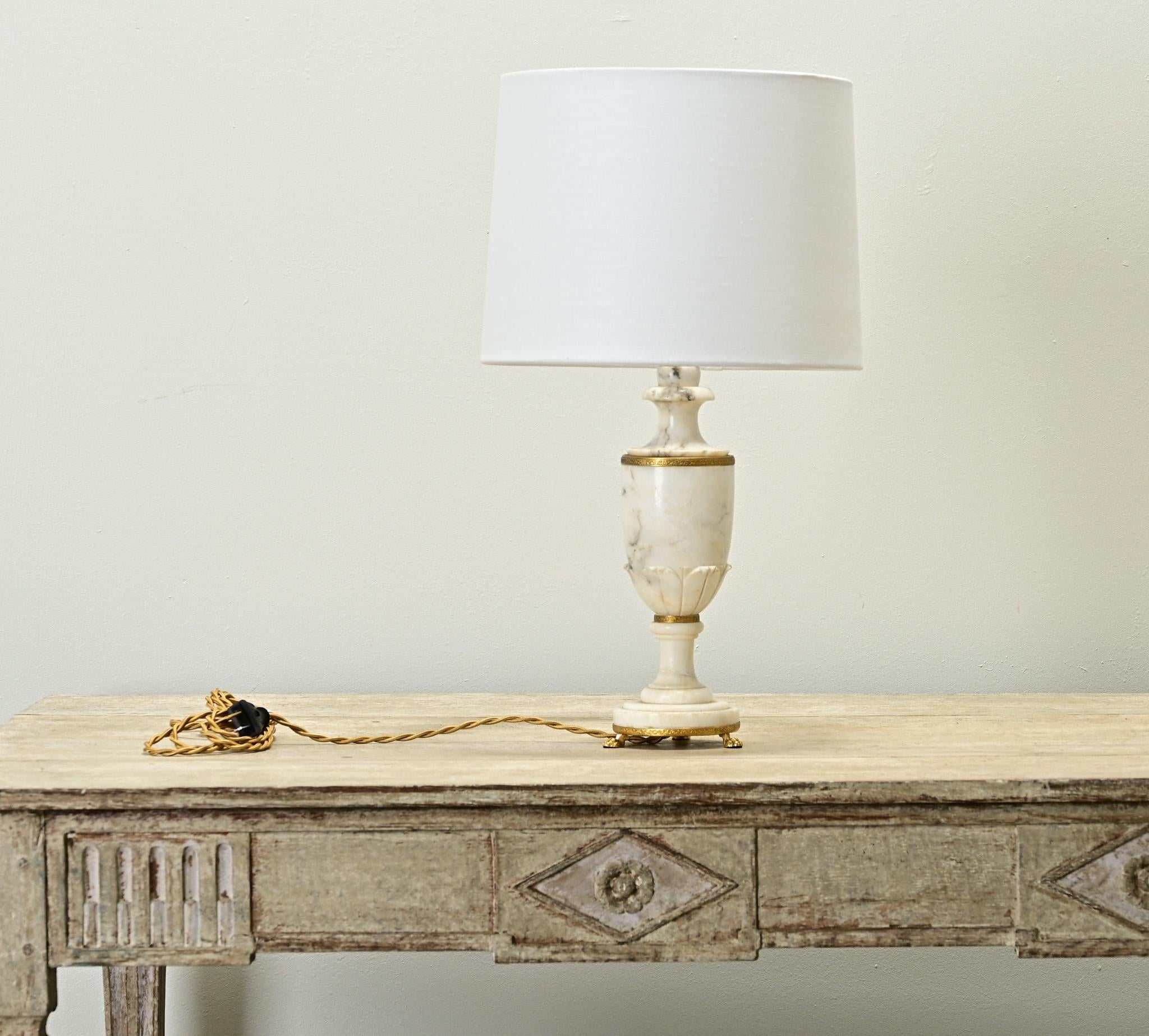 A French Empire carved marble and brass table lamp. This marble lamp is shaped like an urn with gold gilt brass trim and paw feet with a complimentary new linen drum shade. Recently professionally wired for the US using UL listed parts. Be sure to
