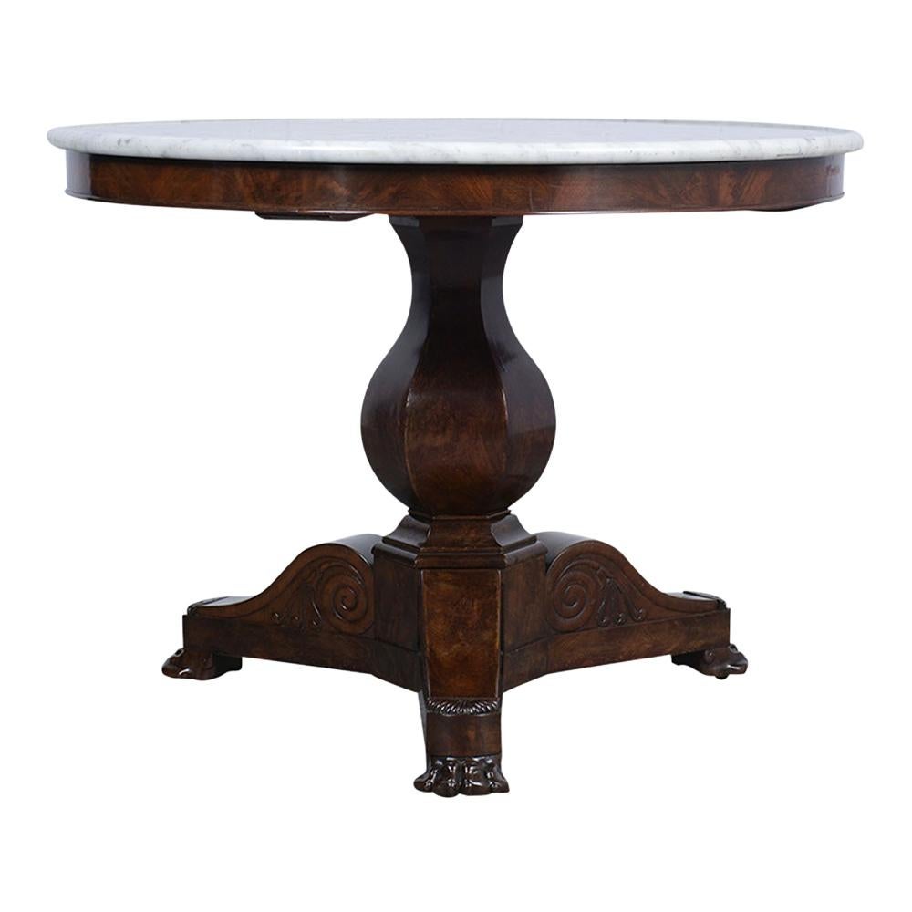 This Antique 1850's French Empire-style Round Center Table is made of solid wood covered in burl veneers stained a rich mahogany color with a patinated finish. The pedestal round table features a bucket shape with ornately carved legs and a white