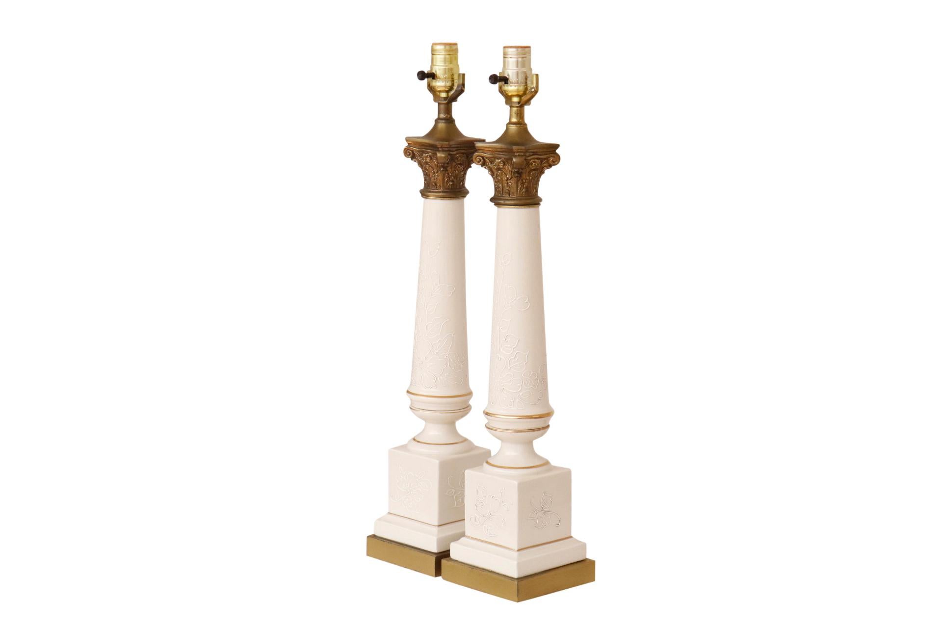 A pair of French Empire style ceramic table lamps by Tyndale. Sleek baluster shaped columns are topped with brass capitals cast with scrolled acanthus leaves. Columns are decorated with a floral motif, mirrored on the block bases. Trimmed with gilt