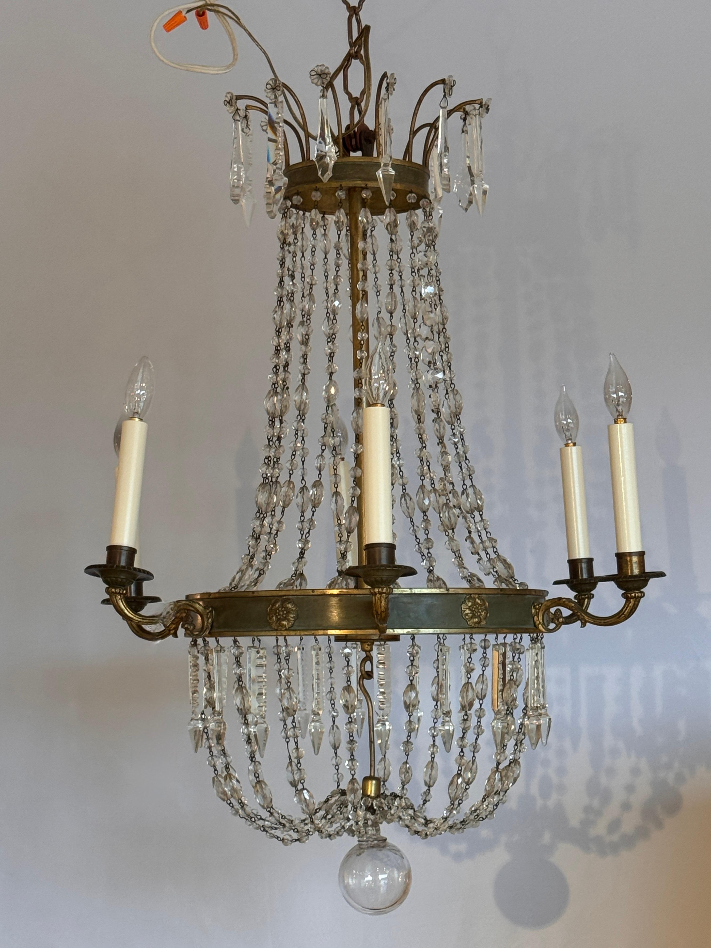 Lampadario in stile Impero francese con struttura in ottone patinato con medaglioni a rosetta e sei bracci a candela. Sospesi, i fasci di perline di cristallo creano una graziosa forma a tenda, che termina con gocce sfaccettate e prismi a lancia. La