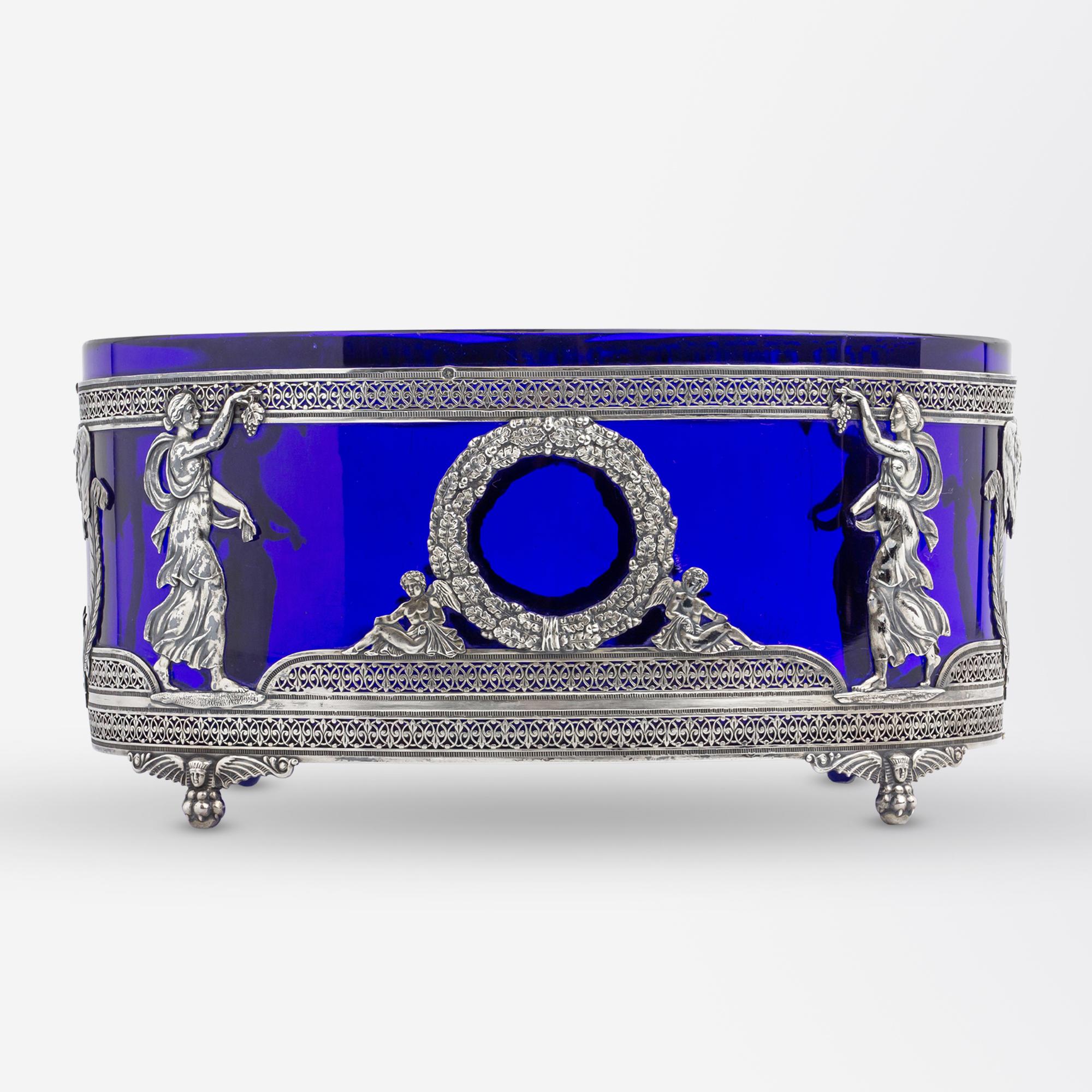 This oval jardiniere which has been crafted from silver and cobalt blue glass dates to France at the end of the 19th Century. The neoclassical jardiniere features Roman or Greek goddesses holding grapes facing toward a central wreath supported by a
